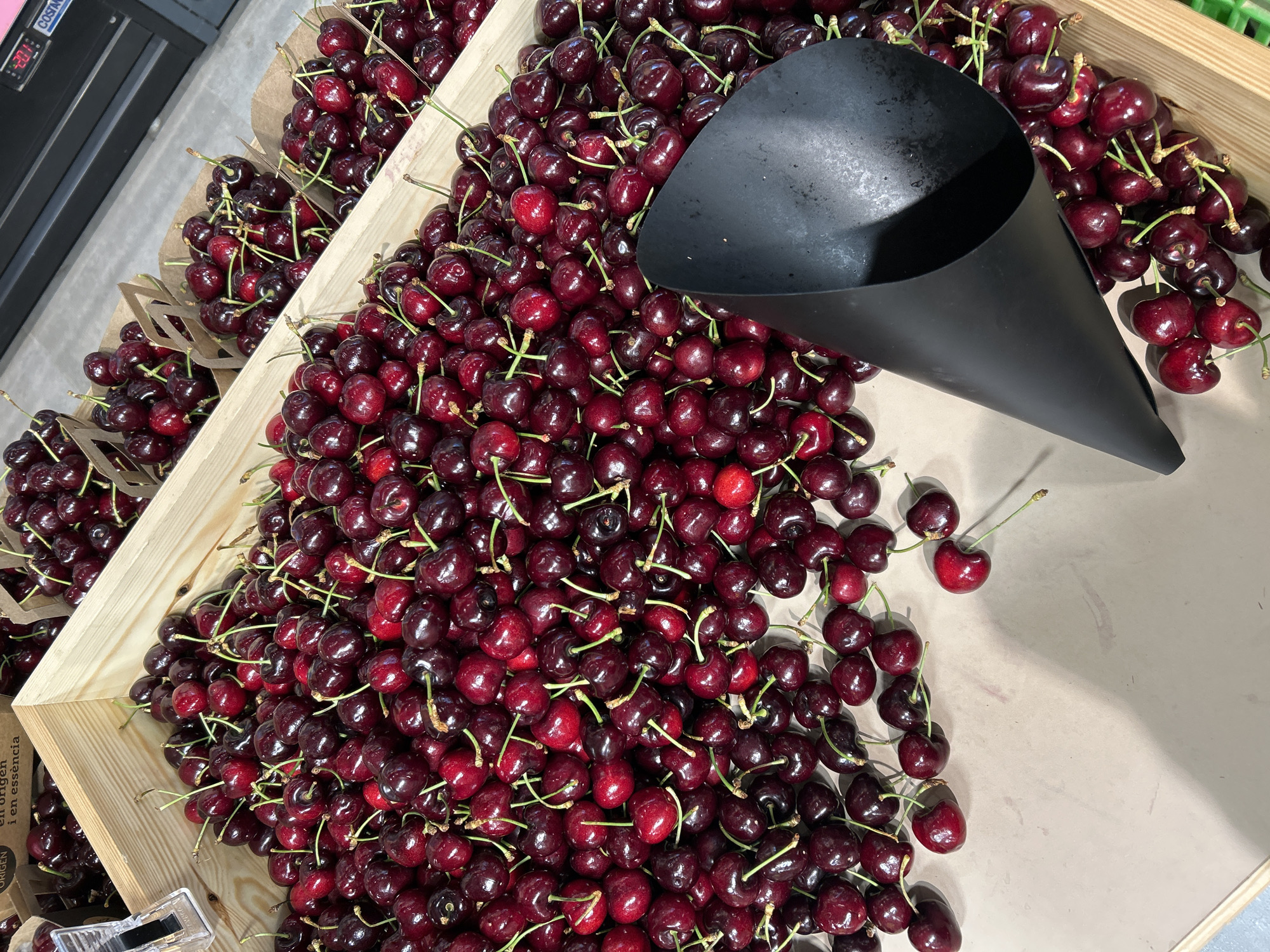 The image shows a close-up, top-down view of several wooden crates filled with ripe, dark red cherries at a market stall in Barcelona, Spain. The cherries are clustered densely, with some spilling out onto the light-colored surface below. A black, cone-shaped scoop rests on top of some of the cherries, partially obscuring them. The lighting suggests an indoor or covered market environment. The background is slightly blurred, showing more crates of cherries and possibly some shelving or display units. The overall impression is of abundance and fresh produce at a commercial point of sale.
