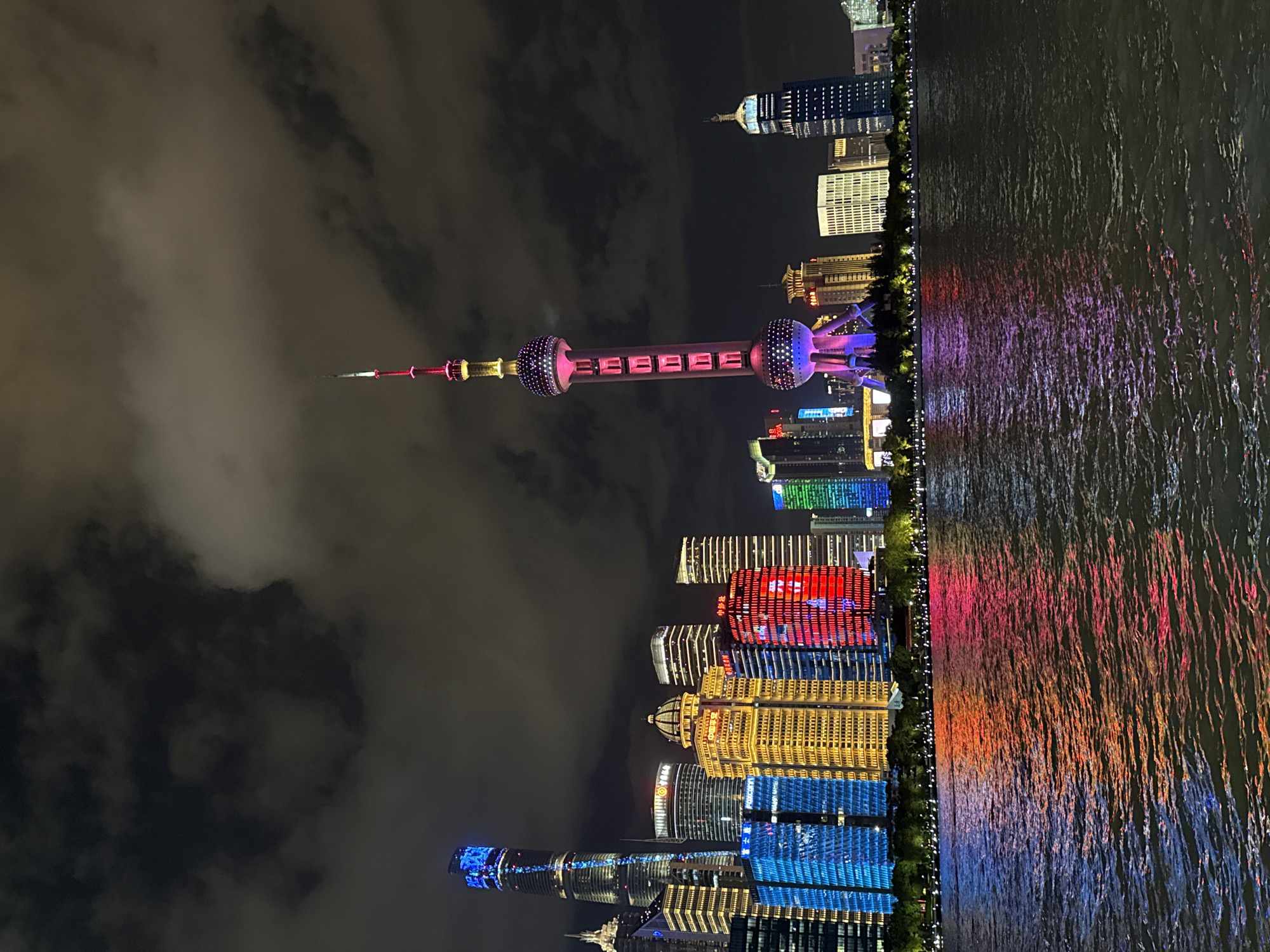 The image depicts a nighttime view of the Pudong skyline in Shanghai, China, as seen from across the Huangpu River. The Oriental Pearl Tower, a prominent landmark, is illuminated with pink and purple lights, with rows of red text displayed vertically on its central shaft. To the left of the tower, several skyscrapers are visible, some lit with vibrant blue, yellow, and red lights. These buildings display geometric patterns and some have Chinese characters on their facades. The foreground is dominated by the dark water of the Huangpu River, reflecting the colorful lights of the city. The sky above is filled with dark, textured clouds, with some lighter areas suggesting moonlight or atmospheric effects. The scene is characterized by artificial illumination and the vast expanse of water. The Hongkou District is not directly depicted in this specific view, as the image focuses on the Pudong side of the river.