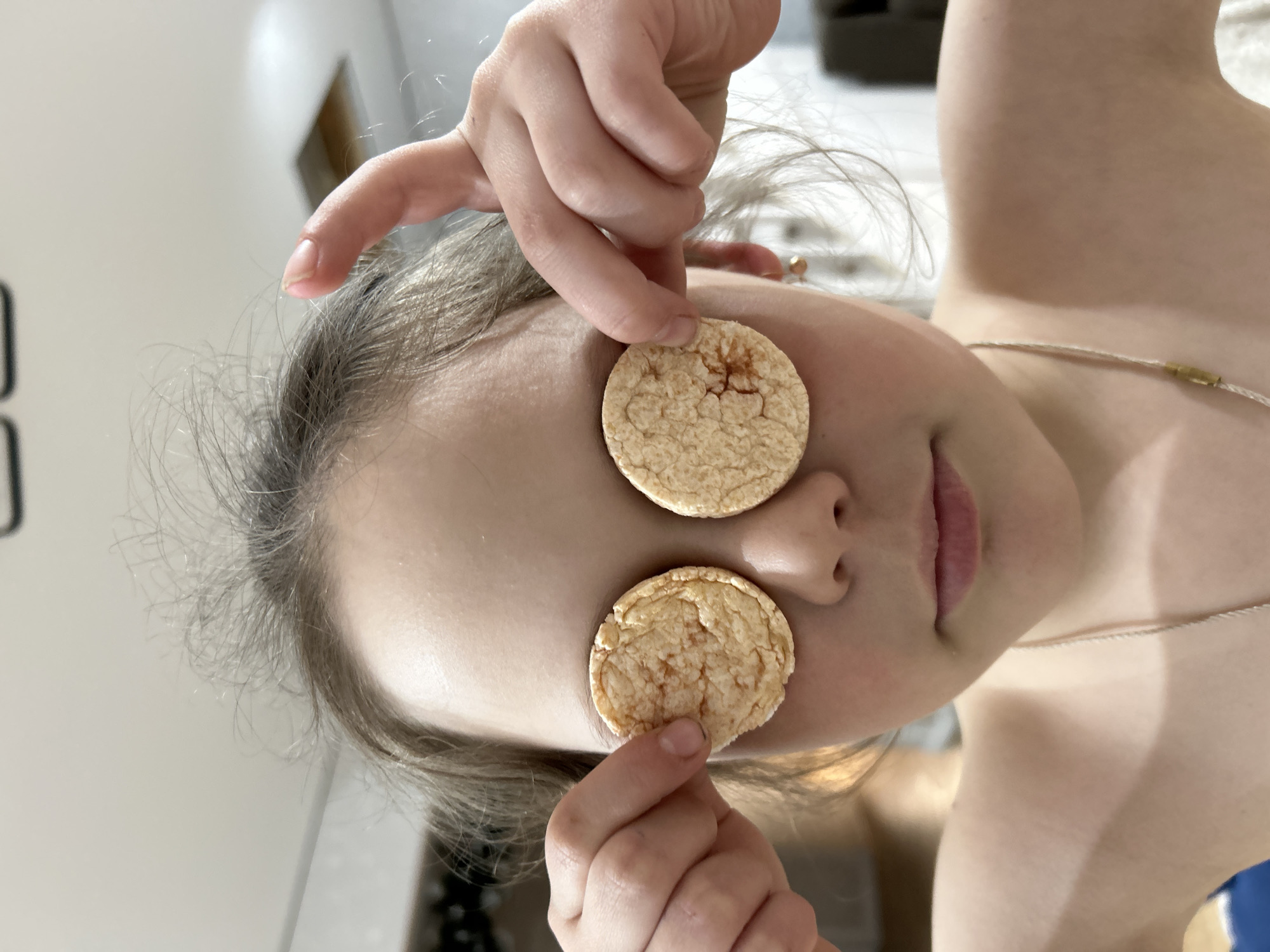 A close-up, rotated image captures a young person, likely a child or pre-teen, playfully covering their eyes with two round, light-colored snack items, possibly rice cakes. The person's head is tilted sideways due to the image orientation. They have fair skin and light-colored hair, some strands appearing messy. Their hands are visible holding the snacks in place, with the fingers and nails clearly seen. A subtle, almost imperceptible smile or playful expression is suggested by the slightly open mouth. Around their neck, a thin, light-colored necklace with a small gold-toned bead is visible. The background is simple and out of focus, suggesting an indoor setting with light-colored walls and hints of a doorframe or furniture, typical of a home environment. The overall mood conveyed is playful and lighthearted. No visible text is present. The event takes place in Chaikovsky, Russia.