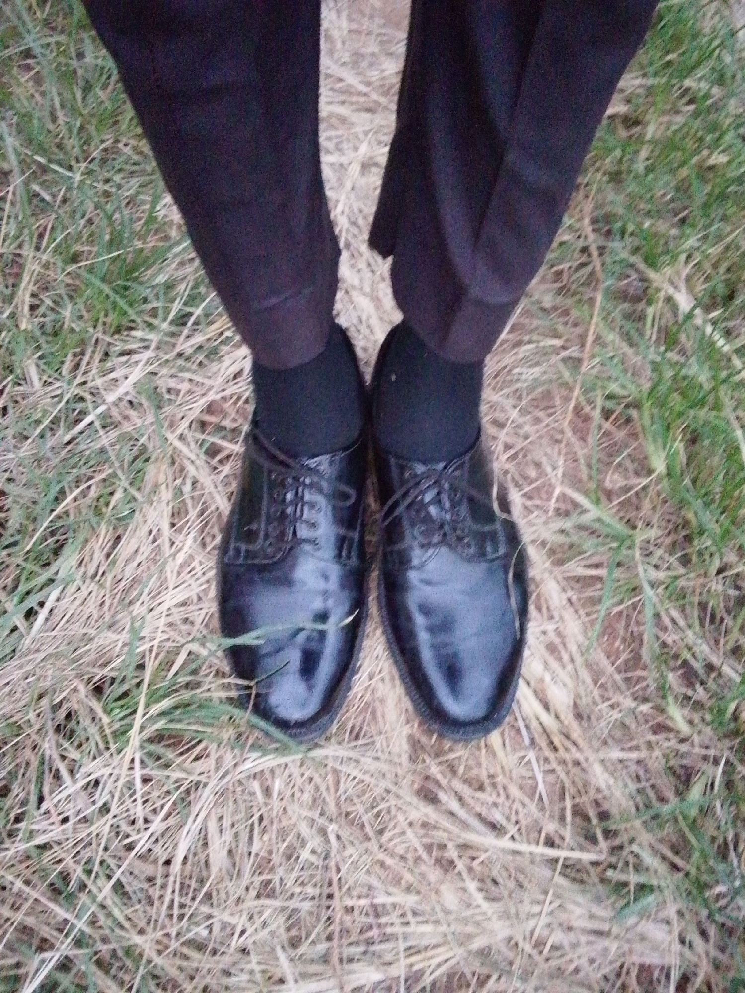 The image shows a close-up view of a person's feet, wearing black dress shoes and dark trousers, standing on a grassy and dry, straw-covered ground. The scene appears to be outdoors, possibly in a rural or semi-rural area. The lighting is natural, suggesting it might be daytime. There is no indication of the city of Serti, Nigeria, or any specific urban elements in the image. The focus is on the footwear and the lower part of the trousers against the natural ground cover.
