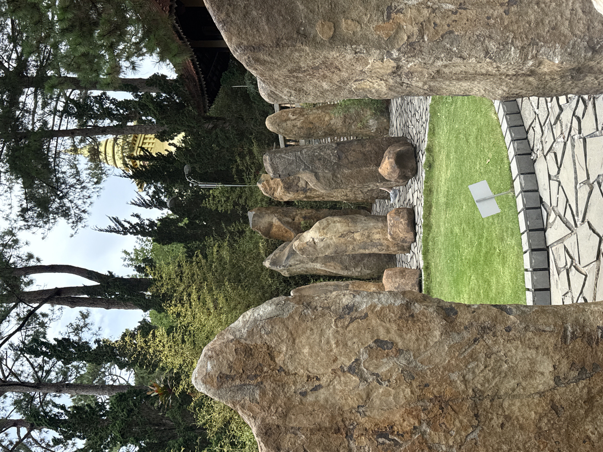 The image shows a stone path or walkway in Da Lat, Vietnam. The path is lined on both sides by irregularly shaped stones of varying sizes and colors, ranging from light tan to darker brown and gray. Between the stone path and the larger stones is a patch of green grass. On one side of the path is a section of patterned stone paving.
In the background, through the spaces between the stones, are trees and foliage, as well as what appears to be a tall golden structure resembling a pagoda. The sky is visible in the upper portion of the scene. The overall impression is of a park or garden setting with a focus on natural stone landscaping.