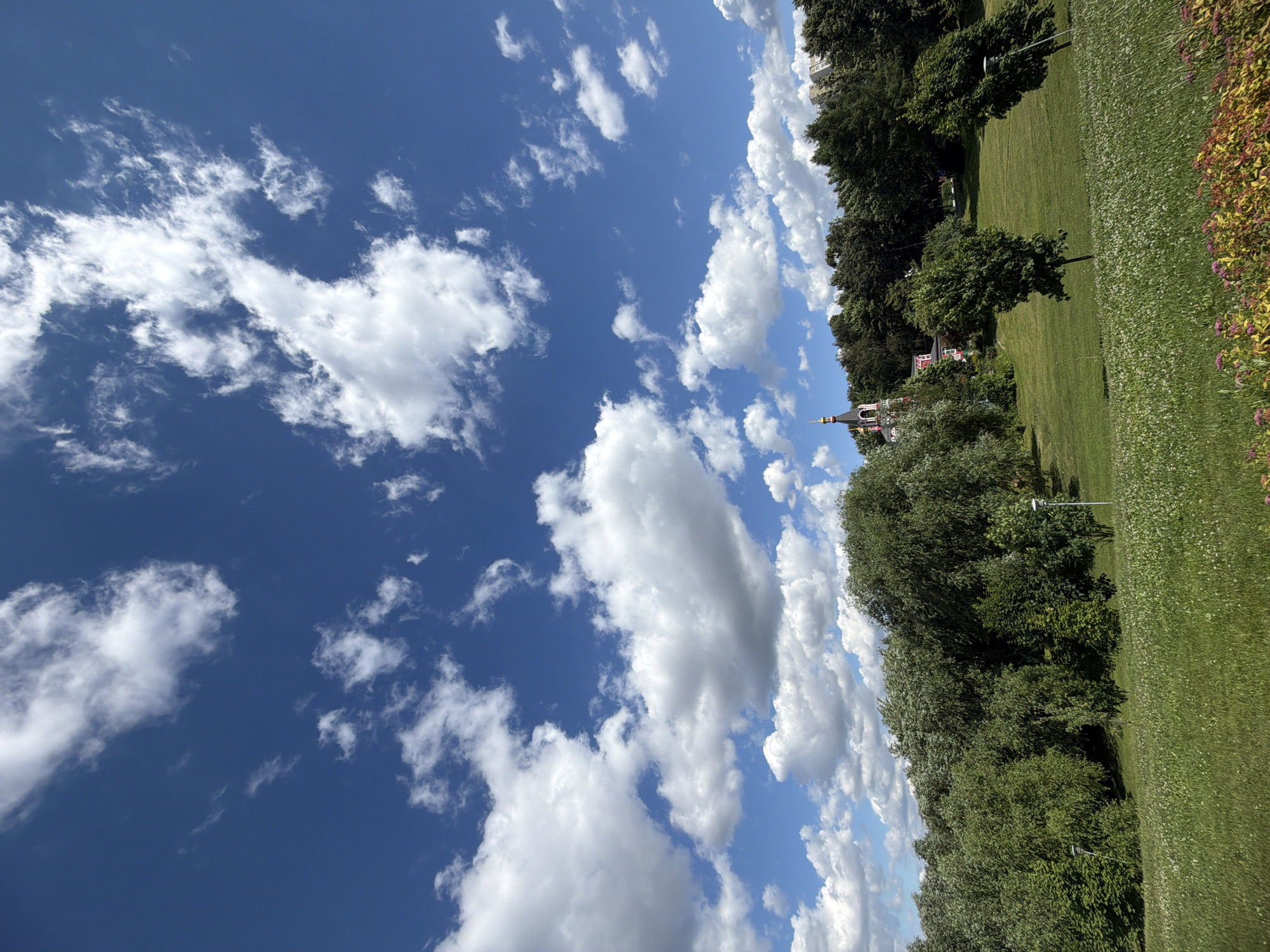 This is a wide-angle, eye-level shot of a park in Moscow, Russia, on a sunny day with a partly cloudy sky. The foreground is a green grassy slope dotted with small white flowers and a patch of pink and yellow flowers at the bottom right. Further up the slope are several trees, some with distinctive lampposts near them. A dense line of trees forms the mid-ground. In the background, a red building with a distinctive gold-domed tower, likely a church, is visible peeking out from behind the trees. A few modern buildings are also visible in the far background on the right side of the frame. The sky is a vibrant blue, filled with fluffy white clouds, suggesting good weather. No people are visible in the scene.