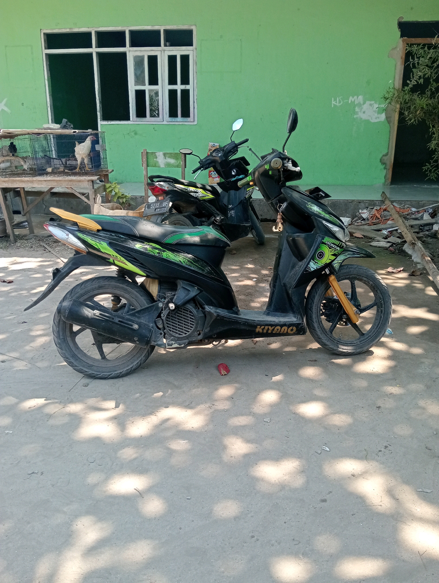 The image depicts a street scene in Pagerungan Kecil, Indonesia, on a sunny day. In the foreground, a black scooter with green accents is parked on a concrete surface. The scooter has the word "KIYANO" printed on its side panel. Behind the scooter, another black scooter is partially visible. The background features a light green building with a large white-framed window. To the left of the building, a wooden table holds a bird cage containing several chickens. The environment is somewhat cluttered, with debris and branches scattered on the ground. Shadows of leaves and branches are cast on the concrete, indicating the presence of trees. The overall impression is one of a typical, somewhat unkempt, outdoor setting in a rural or semi-urban area of Indonesia. There are no people visible in the image.