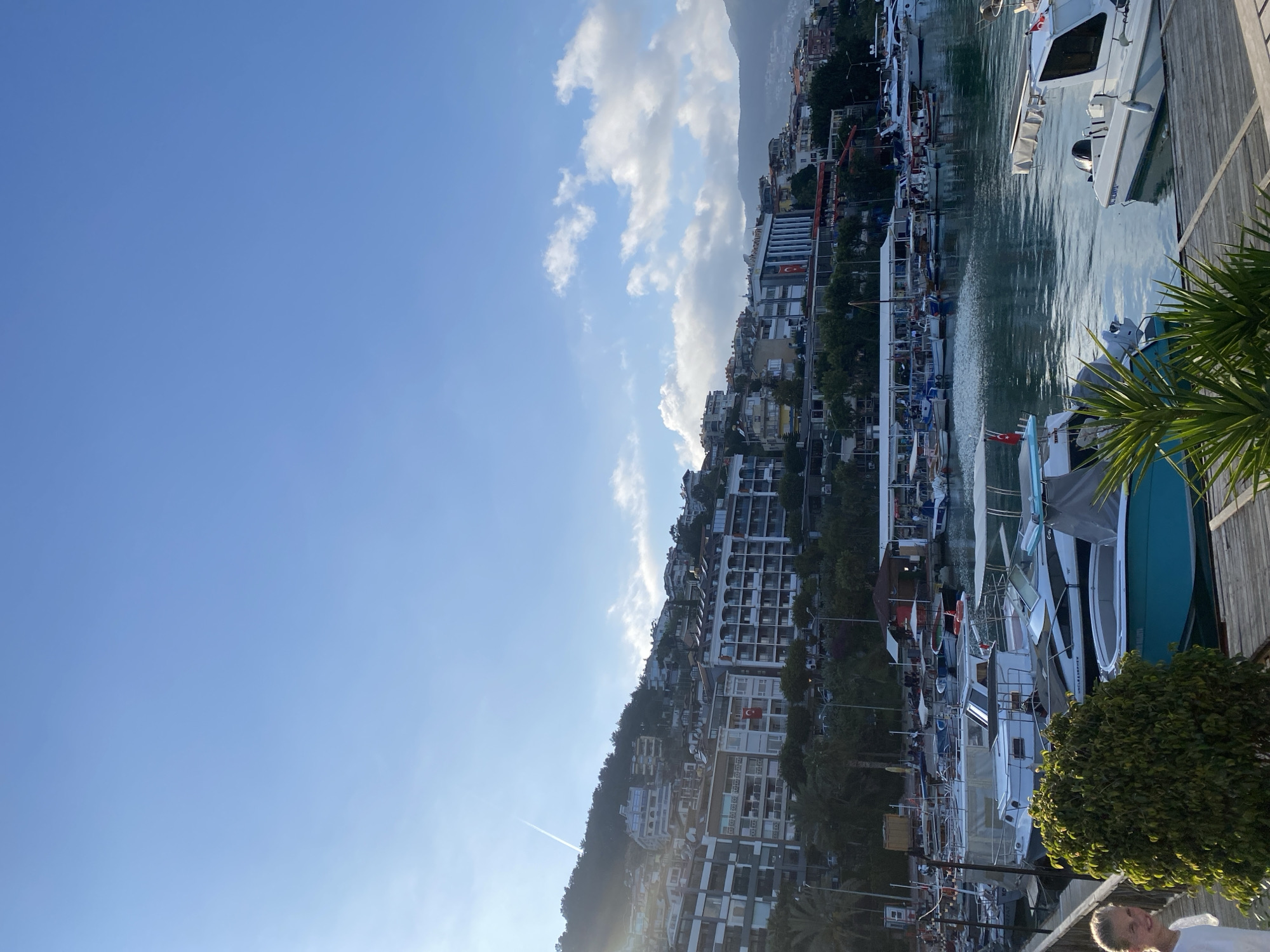 This vertical full-shot image captures a picturesque coastal scene in Alanya, Turkey, viewed from an elevated perspective overlooking a marina. The main subjects include a marina with numerous boats, a town built into a hillside, a distant mountain range, and a clear blue sky.
In the foreground, the top of a person's head and shoulder are visible in the bottom right corner, partially obscured by vibrant green foliage, including palm fronds. The person has light-colored hair and appears to be wearing a white top.
The middle ground is dominated by a marina filled with various boats and yachts, ranging in size and color, predominantly white with some having blue or teal hulls. They are moored along a wooden pier that extends along the right edge of the water. The water surface is relatively calm, reflecting the boats and the sky. Behind the marina, a densely packed town cascades down a lush green hillside. Numerous white and light-colored buildings, likely apartments or hotels, are interspersed with trees and vegetation. A red Turkish flag with a white star and crescent is prominently displayed on one of these buildings, providing a clear location cue.
In the background, a majestic mountain range rises, its peaks partially veiled by scattered white clouds. The sky above is a brilliant, clear blue with additional white, fluffy clouds on the right side. A faint contrail from an aircraft is visible stretching across the lower left part of the sky.
The time of day appears to be late afternoon or early morning, with the sun positioned low, creating a warm glow and a subtle sunburst effect in the lower-left portion of the image. The weather is sunny with some scattered clouds. The overall scene conveys a serene and beautiful Mediterranean setting.