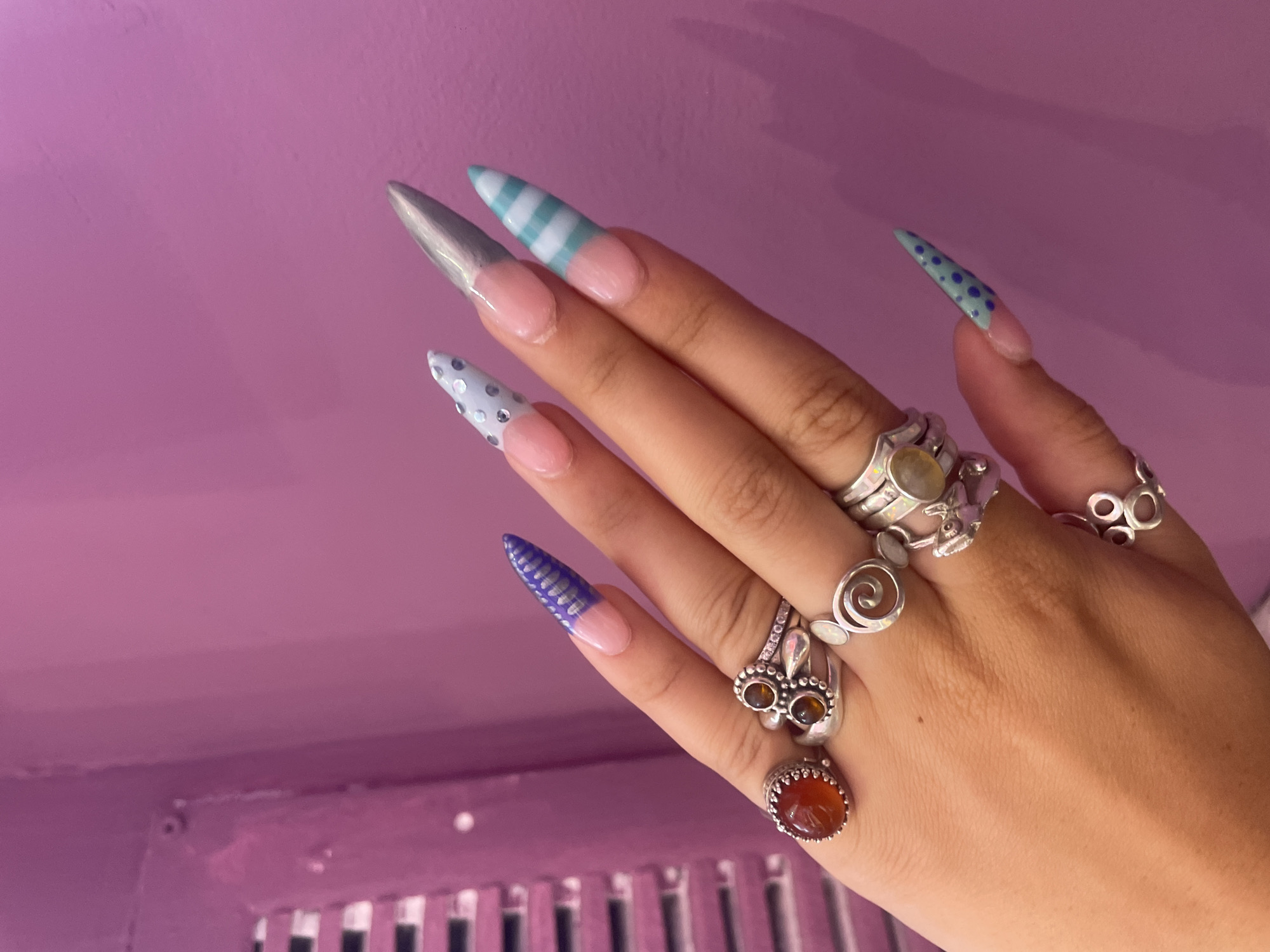 The image shows a close-up of a person's left hand, adorned with several rings and long, stiletto-shaped acrylic nails. The nails feature various designs, including a purple wavy pattern, light blue with silver polka dots, metallic silver, light blue and white stripes, and light green with dark blue polka dots. The background is a solid, vibrant pink wall, with a hint of a gridded, possibly ventilation, structure on the left side of the frame. The lighting suggests an indoor setting. The exact location in Raanana, Israel, is not discernible from the visual information provided.