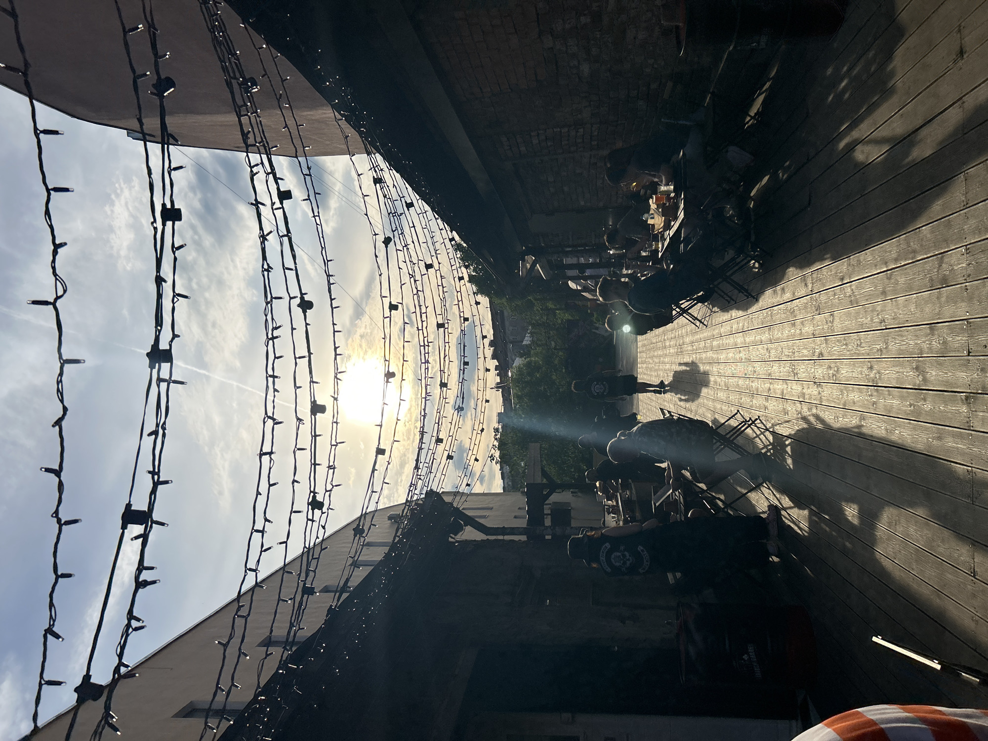 This image depicts an outdoor alleyway or courtyard cafe in Saint Petersburg, Russia, at late afternoon or early evening, characterized by strong backlighting from a low sun. Numerous parallel strings of small, white decorative lights are suspended overhead between two buildings that frame the space. The building on the left features a light-colored facade with visible windows, while the building on the right has dark brickwork. The ground is covered by a wooden deck, showing pronounced sun glare and long, dark shadows cast by people and objects.
Multiple individuals are present, most of whom are seated at dark, foldable tables and chairs arranged along both sides of the alley. They are engaged in social interaction, consuming beverages and food from glasses and bottles on their tables. On the left side, one person wears a dark vest with a skull and crossbones emblem visible on the back. In the central mid-ground, a lone figure walks away from the viewer down the sunlit wooden path, casting a distinct long shadow. Scattered greenery and distant urban structures are visible in the background beneath a partly cloudy sky, which also shows a faint contrail in the upper left. Dark barrels, one labeled "anarchy barrel," are positioned near the seating areas. A portion of a person's arm, clad in an orange and white striped shirt, is visible in the bottom left foreground.