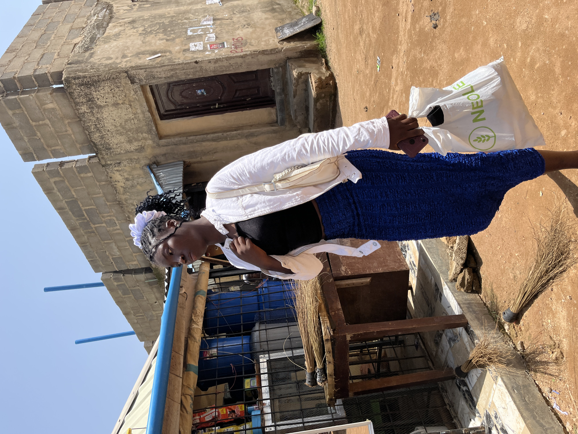 A young Black woman walks away from the camera on a dusty, unpaved street in Jos, Nigeria. She is wearing a white jacket over a black tank top and a textured blue skirt. She carries a white plastic bag with a green logo and a cell phone in her right hand. Behind her, a storefront displays various goods behind a metal grate. The building behind the storefront is made of concrete blocks and has a weathered, tan facade. Two brooms lie on the ground in front of the storefront. A thin layer of dust covers the ground and the lower parts of the buildings. The sky is clear and bright blue.