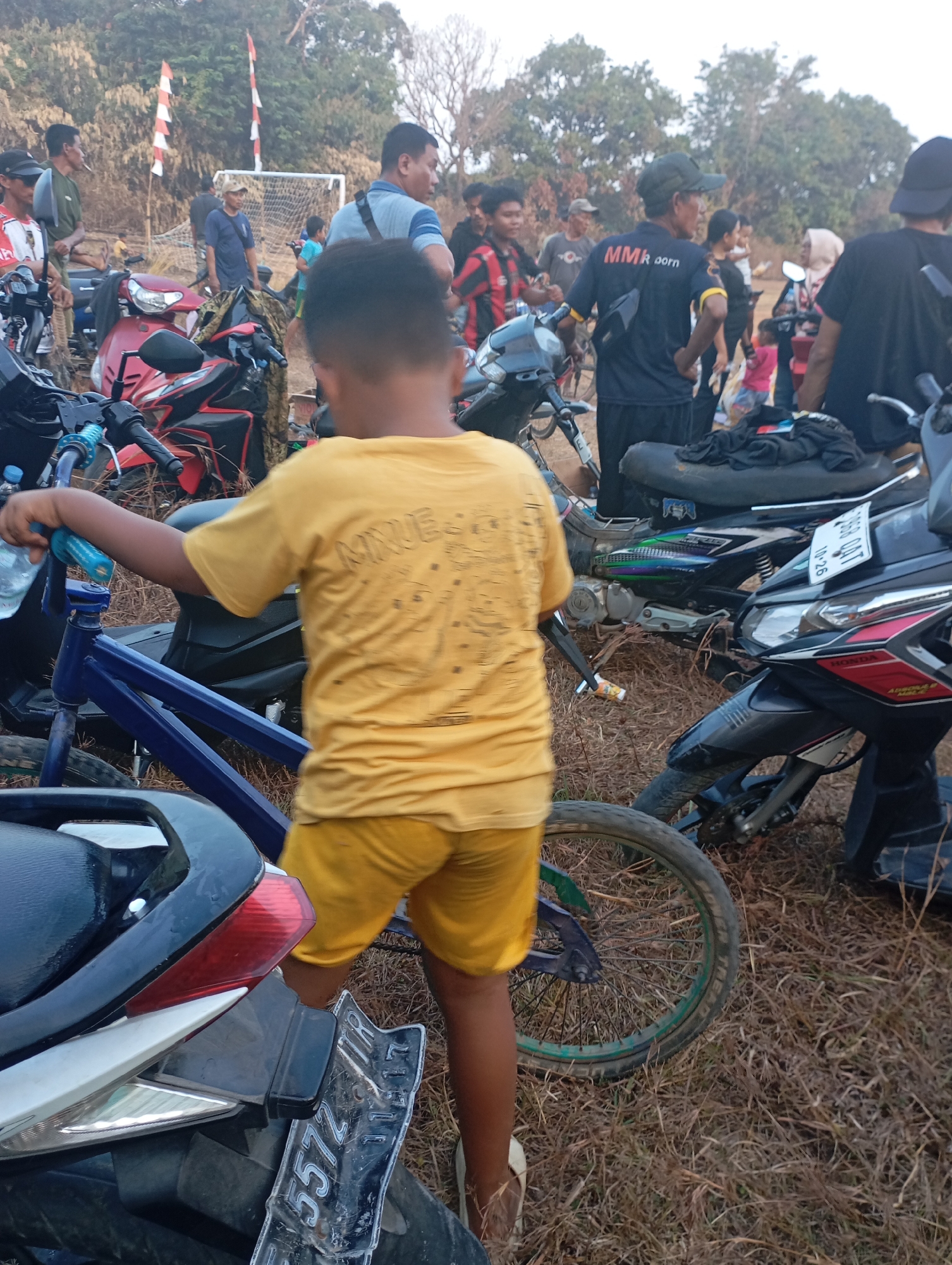 In Kecamatan Haurgeulis, Indonesia, a group of people are gathered outdoors, possibly for an event. The ground is covered in dry grass and twigs, suggesting a rural or undeveloped area. Several motorcycles are parked, indicating a mode of transportation for the attendees. A young boy, wearing a yellow t-shirt and shorts, is the central focus of the image, standing near a blue bicycle. In the background, a soccer goal is visible, hinting at a sporting activity or a recreational gathering. The sky is overcast, and the lighting suggests it's either early morning or late afternoon. The overall atmosphere appears casual and communal.
