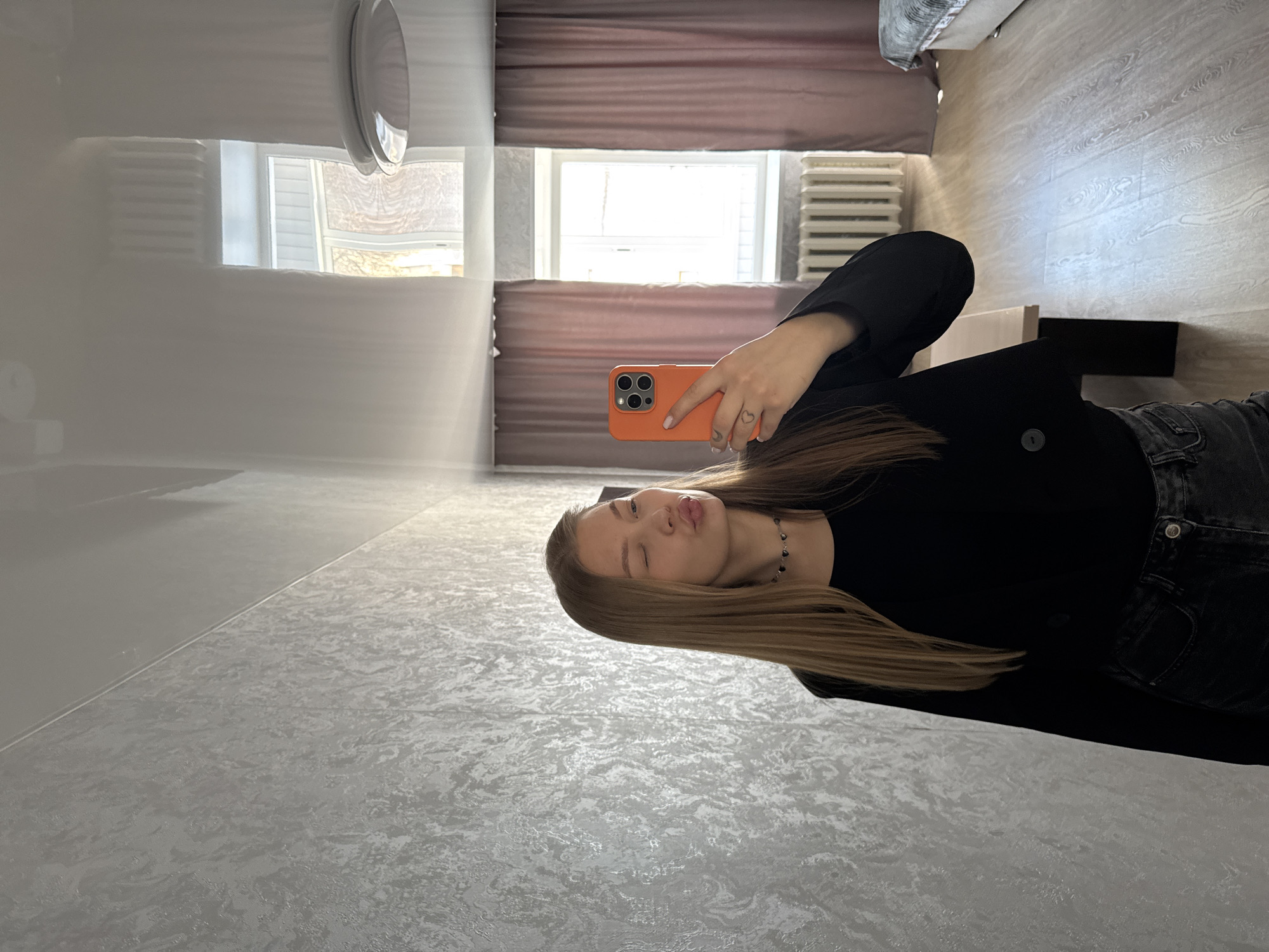 A young woman with long, straight, light brown hair is taking a mirror selfie in an indoor room, likely in Chaikovsky, Russia. She is wearing a black long-sleeved top and dark jeans, accessorized with a dark beaded choker necklace. She holds an orange smartphone in her right hand, which shows a small heart tattoo on her ring finger. Her expression is playful, with pursed lips as if blowing a kiss and one eye winking.
The setting appears to be a bedroom or living space. The room features a light-colored wooden floor and textured light gray walls. A window with a white frame allows bright natural light into the room, suggesting it is daytime. Below the window, a white radiator is visible, and light brown curtains flank the window. The glossy white ceiling reflects parts of the room, including a circular light fixture. A bed or sofa is also visible in the background reflection. The scene is well-lit, and the woman's expression conveys a confident or playful mood. There is no visible text in the image.