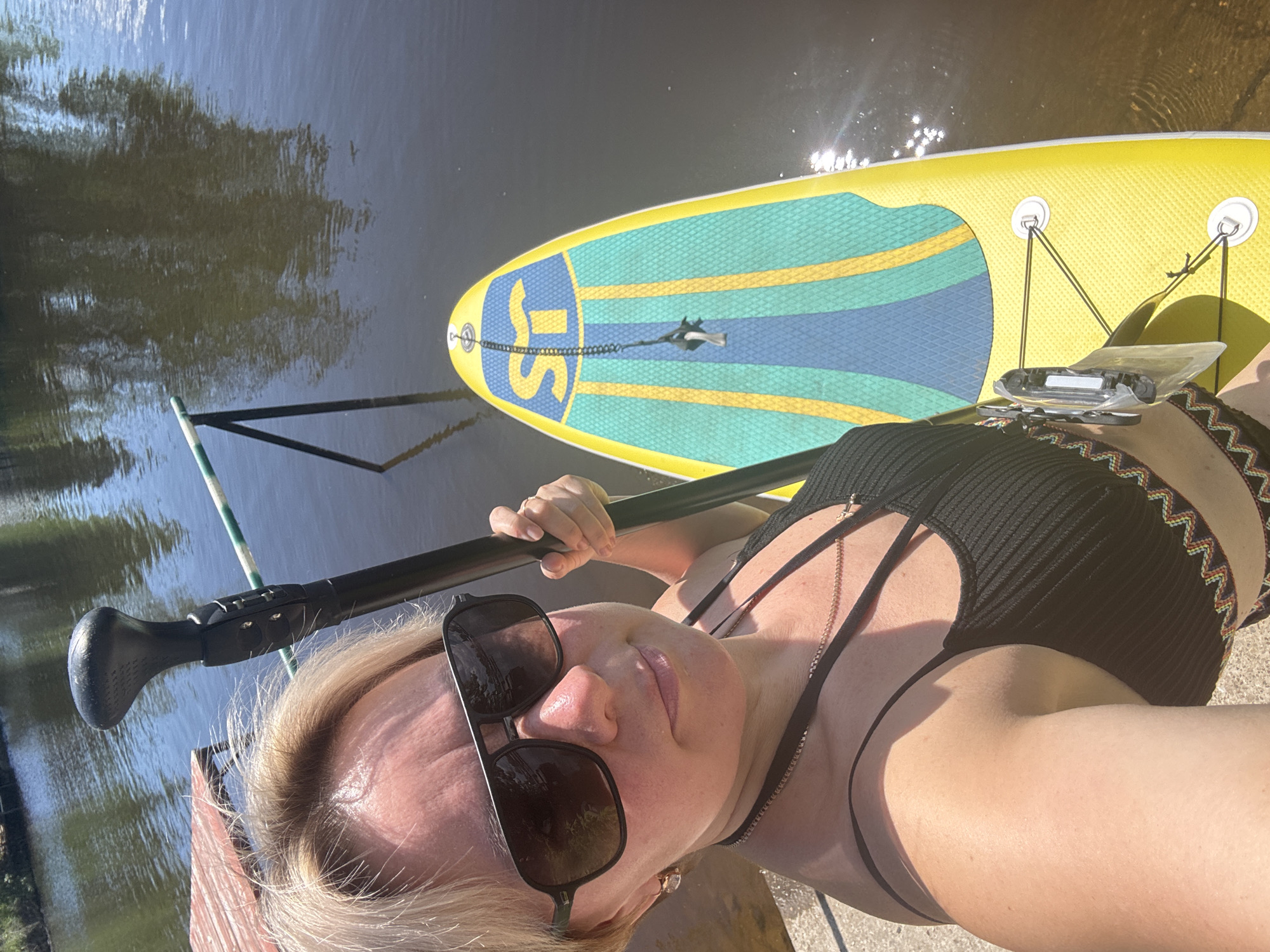 A woman with blonde hair and sunglasses is holding a paddleboard paddle, standing next to a yellow and blue paddleboard. She is wearing a black bikini top with a patterned trim. The paddleboard is in the water, which reflects trees. A wooden dock is visible on the left side of the image. The scene appears to be a body of water, possibly a river or lake, in a natural environment.