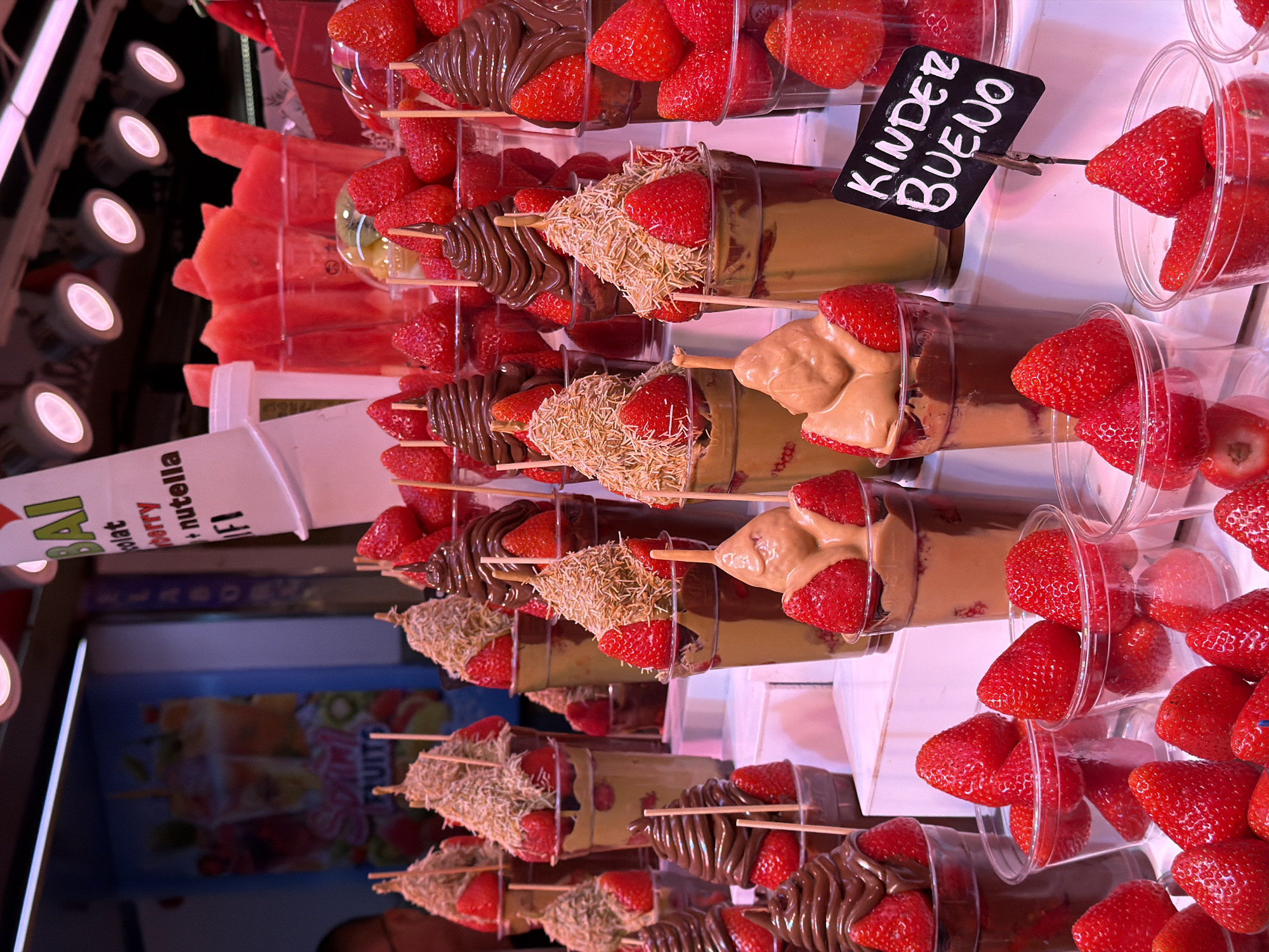 The image depicts a vibrant display of desserts, likely in a market stall, featuring an abundance of fresh strawberries dipped in chocolate and other sweet toppings. Several clear plastic cups contain layered desserts with strawberries, some topped with a swirl of chocolate or a peanut butter-like cream. There are also cups filled with cut watermelon and what appear to be fruit smoothies in the background. A sign that reads "Kinder Bueno" is visible in the foreground, suggesting a connection to a popular chocolate bar. The overall scene suggests a dessert vendor, possibly in a busy urban area. While specific clues to the exact location in Barcelona are not evident from the image itself, such stalls are commonly found in popular tourist areas and markets throughout the city. The lighting is bright, suggesting an indoor or covered setting.