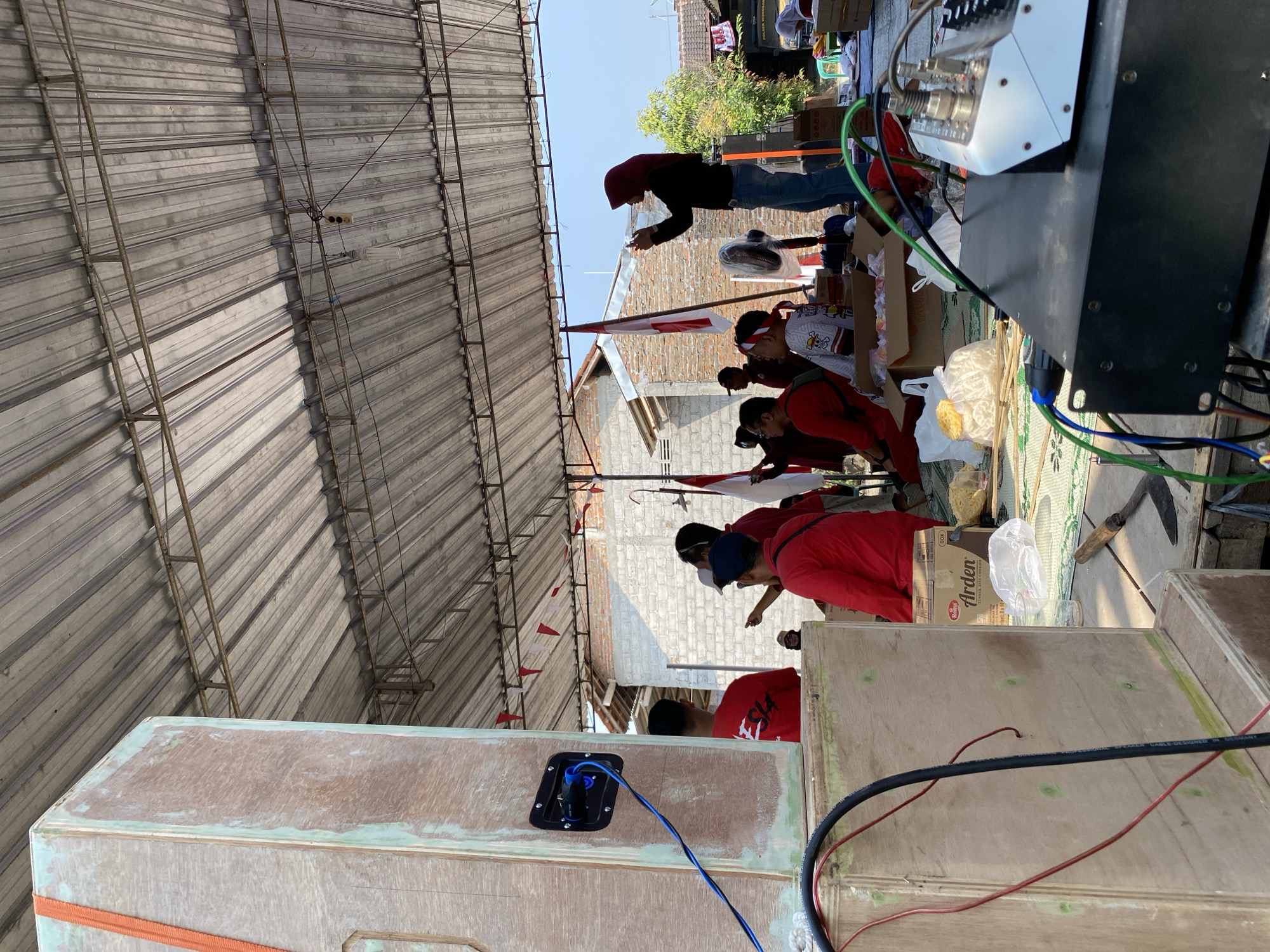 This image captures an outdoor gathering in Wonosari, Indonesia, under a corrugated metal roof supported by metal beams. In the foreground, a large wooden speaker is visible on the left, with cables connected to it. A black audio mixer sits in the lower right corner, with several cables plugged into it. A group of people, mostly dressed in red, are gathered in the middle ground, some standing and some sitting. One person in the background, wearing a red hijab, is holding a phone and appears to be taking a photo or video. Indonesian flags are present in the scene, suggesting a patriotic or celebratory event. The environment appears to be a semi-urban or rural setting, with brick buildings and trees visible in the background. The overall atmosphere seems informal and communal.