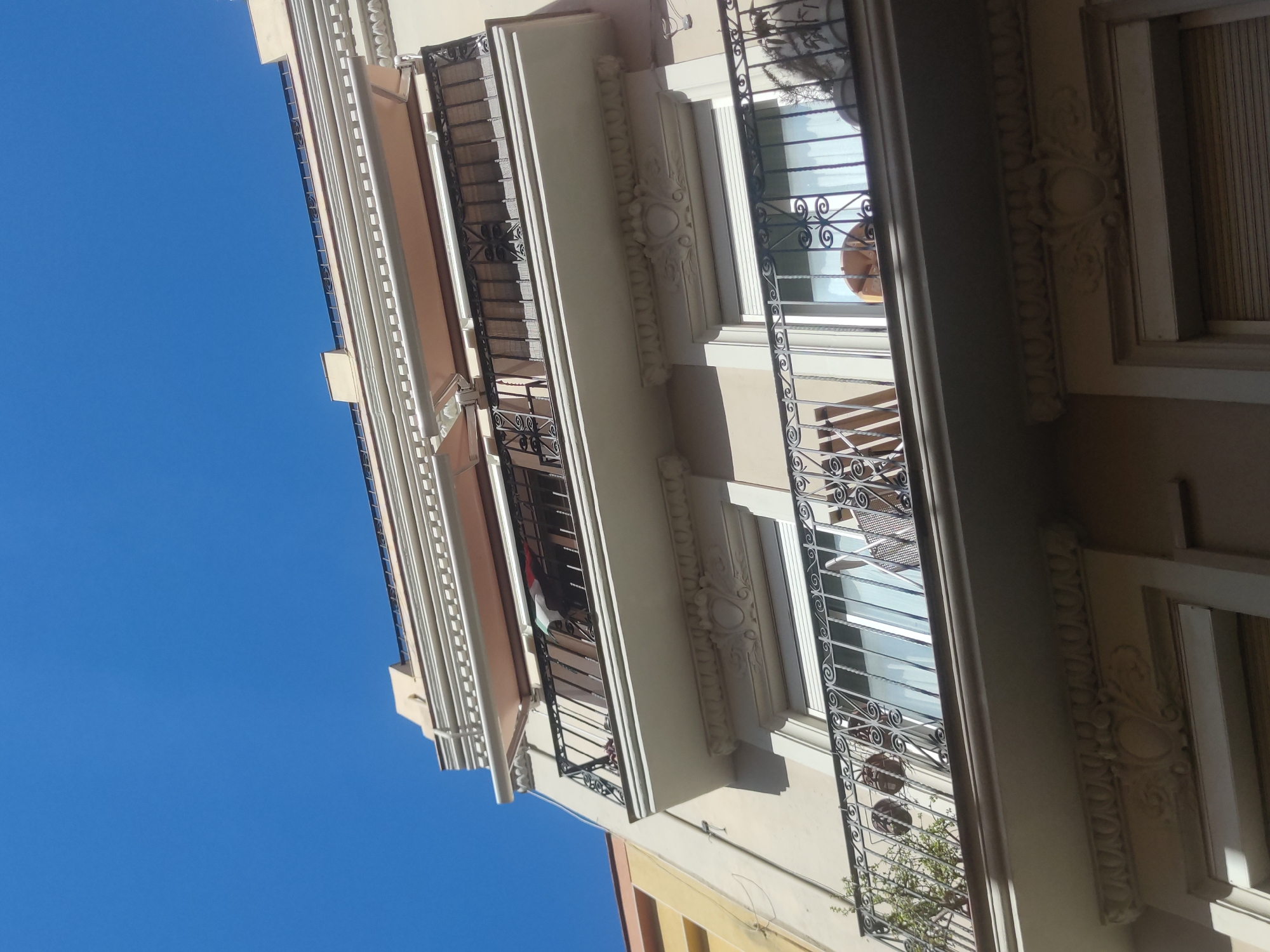 A low-angle shot showcases the exterior of a multi-story building in an urban environment. The building's façade features architectural details such as decorative moldings, wrought iron balconies, and awning-shaded windows. The building's color scheme includes beige, white, and tan tones, contrasting with the clear blue sky visible behind it. The balconies hold potted plants, adding a touch of greenery to the urban scene.