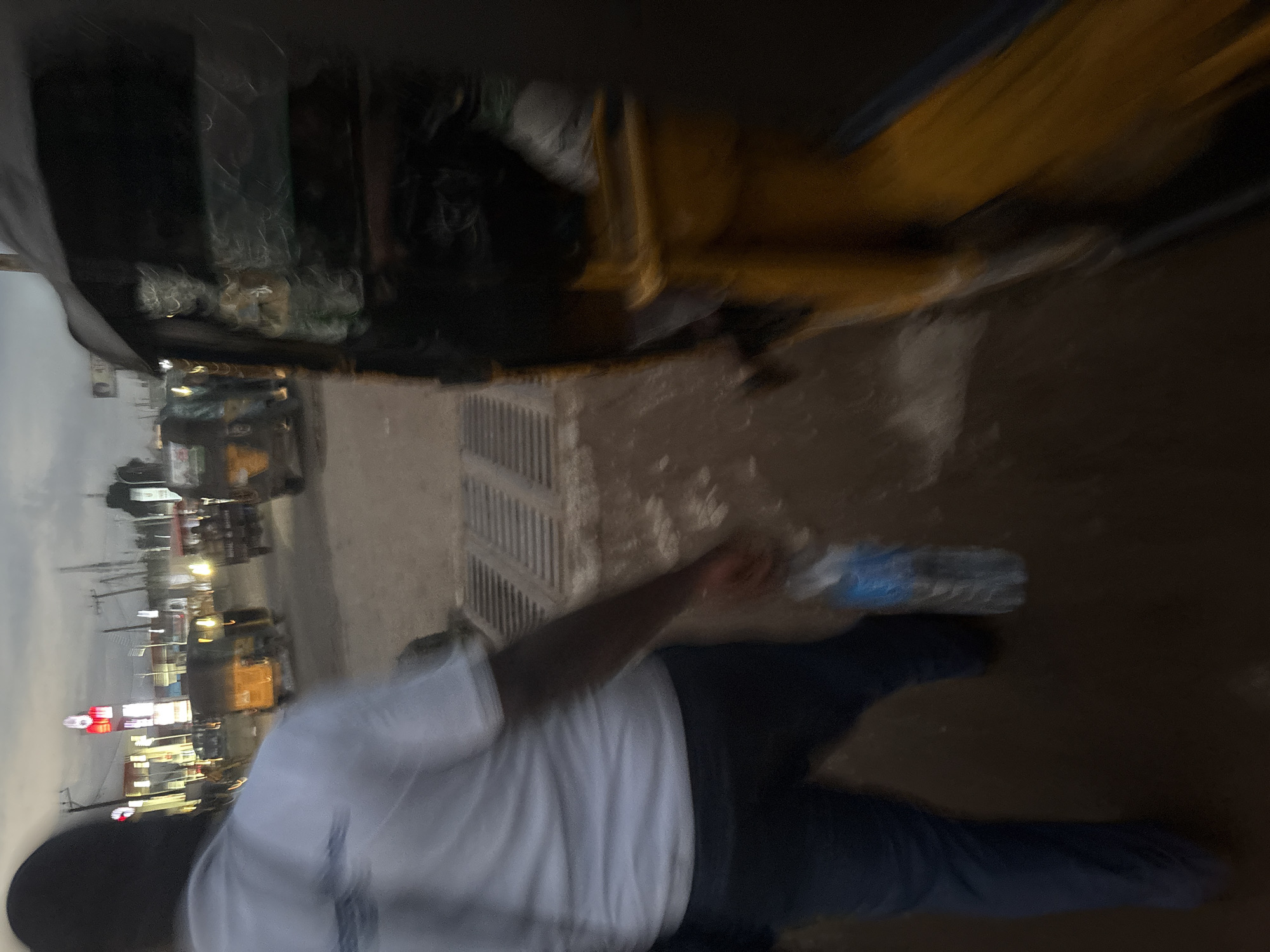 The image depicts a street scene in Onitsha, Nigeria, likely during twilight or overcast conditions, given the muted lighting and sky. In the foreground, a person is walking away from the camera, wearing a white t-shirt and dark pants. They are holding a transparent plastic bottle of water in their right hand. The person's face is not visible.
To the right of the person, a yellow auto-rickshaw, a common mode of transportation in Nigeria, is partially visible, its front section extending out of frame. The auto-rickshaw appears to be stationary or moving slowly. In the background, other auto-rickshaws and vehicles are present on the street. Buildings and illuminated signs are visible in the distance, suggesting an urban environment with commercial activity. The road surface appears to be paved, with some visible drainage grates in the foreground. The overall impression is of a busy street in a Nigerian city.