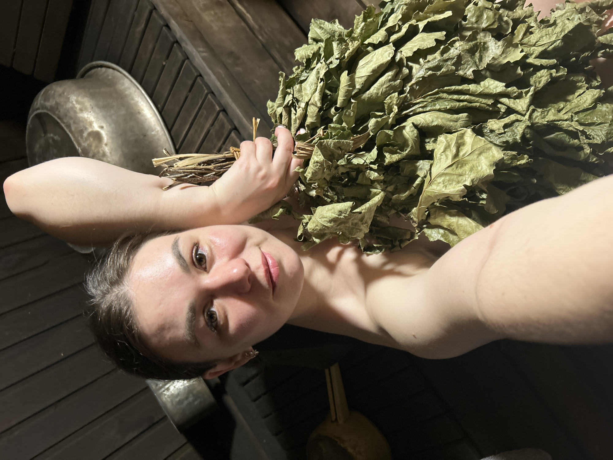 A young woman takes a selfie in what appears to be a traditional banya (Russian sauna). She is pictured from the upper chest upwards, with her body angled slightly and her head tilted. Her skin looks flushed, indicative of the heat within the banya. She has brown hair, which appears to be pulled back, and her brown eyes look directly at the camera with a calm, relaxed expression and a subtle smile.
She holds a large bundle of dried, green-brown leaves, commonly known as a *venik* (besom), in her right hand. The *venik*, likely made from oak or birch branches, is positioned over her left shoulder and chest, partially obscuring her bare shoulder.
The background consists of dark, horizontal wooden planks, characteristic of sauna interiors. To the left, a large, rounded metallic basin or bucket is visible, reflecting some light. In the lower right corner, another smaller metallic object, possibly a ladle or scoop, can be seen. The lighting is dim and warm, highlighting the woman's face and the texture of the dried leaves. The scene conveys a moment of relaxation during a banya session. No text is visible in the image.