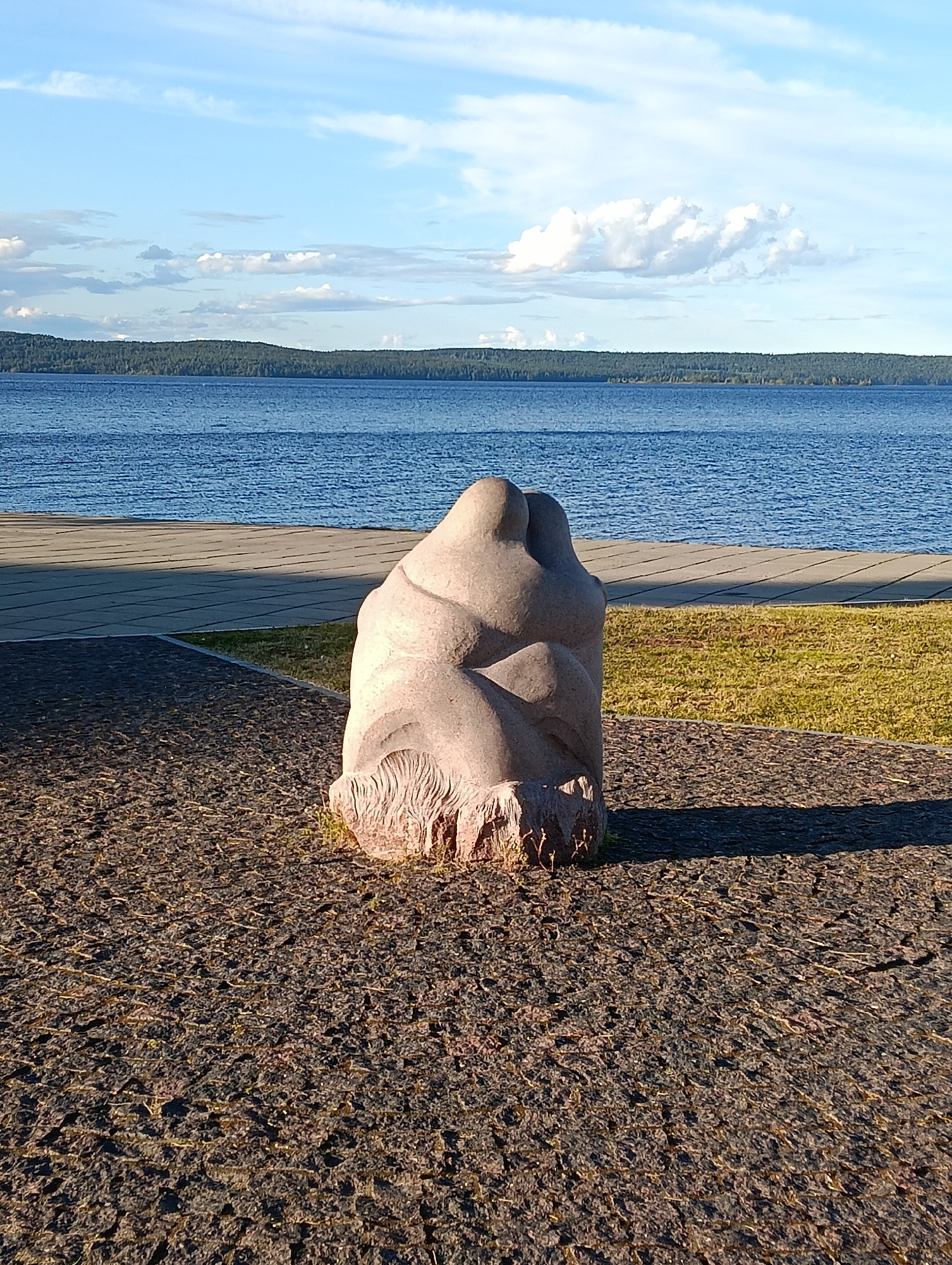 A stone sculpture of two figures embracing is situated on a dark, gravelly surface. The sculpture is positioned in front of a body of water, likely Lake Onega, with a line of trees visible on the far shore. A paved walkway runs parallel to the water, and a grassy area is to the right of the sculpture. The sky above is blue with scattered white clouds. The sculpture is located in the city of Petrozavodsk, Russia, specifically on the Onega Embankment.