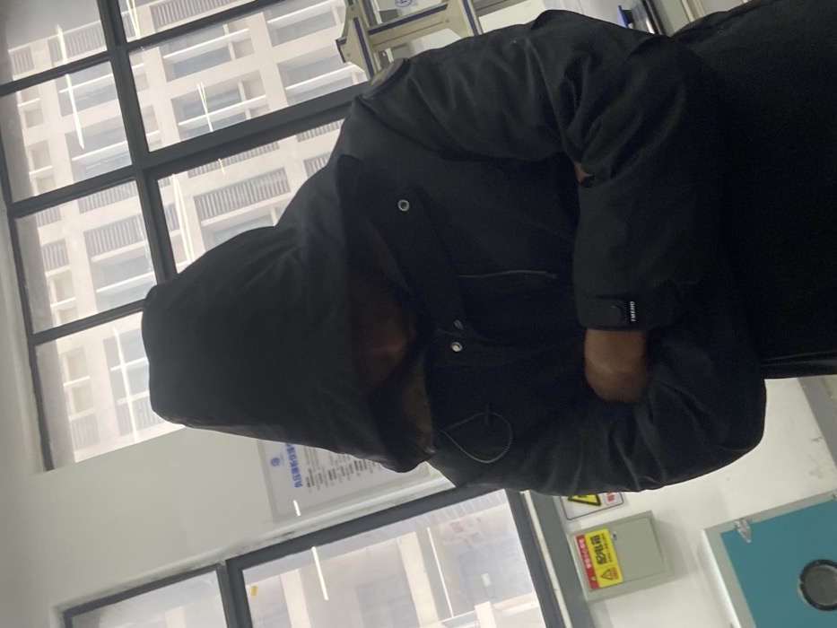 A person wearing a black hooded jacket is standing inside a building. The hood obscures their face, and the jacket appears puffy. A large window with a grid pattern is visible behind the person, showing an out-of-focus building exterior. A sign is partially visible behind the person, and a yellow electrical box is seen in the lower right corner of the frame.