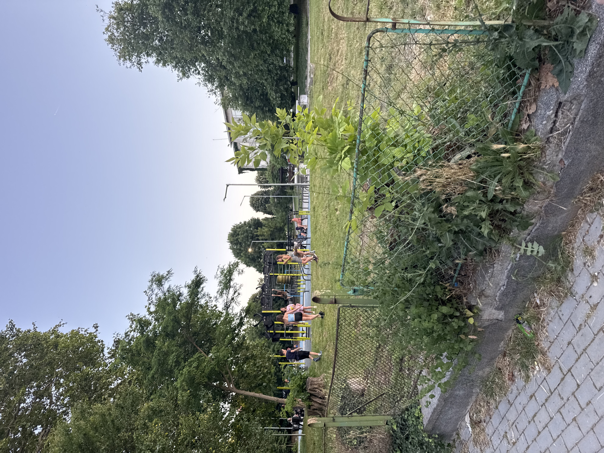 This image depicts an outdoor area in Varna, Bulgaria, featuring a park or recreational space. In the foreground, a weathered green metal fence runs horizontally across the frame, partially overgrown with grass and weeds. Beyond the fence, a grassy lawn is visible, dotted with trees.
Several people are present in the middle ground. A group of young people, appearing to be teenagers or young adults, are gathered near some outdoor exercise equipment. One young male, wearing a dark t-shirt and shorts, is walking away from the camera. Three young females are standing together, one in a blue top and shorts, another in a pink top, and a third in a light-colored dress. Another young female in a light-colored dress is observed further to the right, near the exercise equipment. In the background, more people are seated on benches and around what appears to be a ping pong table.
The environment is characterized by lush green trees, a clear sky, and well-maintained park facilities, including lamp posts and the aforementioned exercise equipment. The presence of both recreational equipment and people suggests a public gathering space designed for leisure and physical activity. The overall scene conveys a sense of community and outdoor enjoyment.
