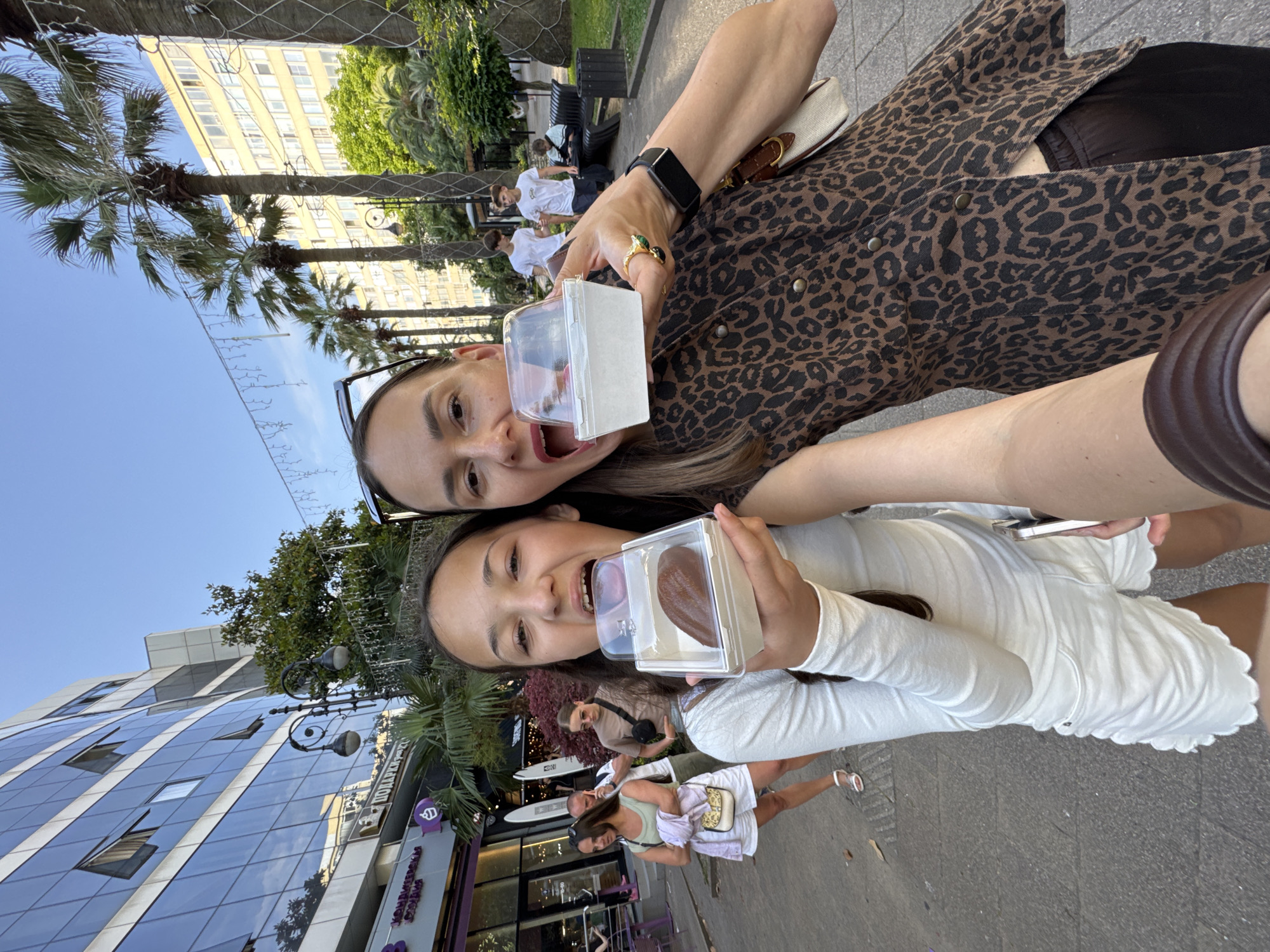 A selfie captures a woman and a girl standing on a paved sidewalk in front of a modern building with a glass facade. Both are holding up clear plastic containers with treats, and their mouths are open as if about to eat them. The girl, on the left, wears a white long-sleeved shirt and white shorts with a scalloped hem. She has dark hair and is holding a dark brown, oval-shaped item in her container. The woman, on the right, has dark hair and is wearing a brown leopard-print top. She is holding a white container. Behind them, palm trees line the street, and other people can be seen walking and sitting on a bench in a grassy area. The sky is clear and blue. The scene suggests a casual outing in a city environment, possibly a tourist area. The presence of palm trees and the architectural style of the buildings are consistent with locations like Sochi, Russia, a city known for its subtropical climate and resort atmosphere.