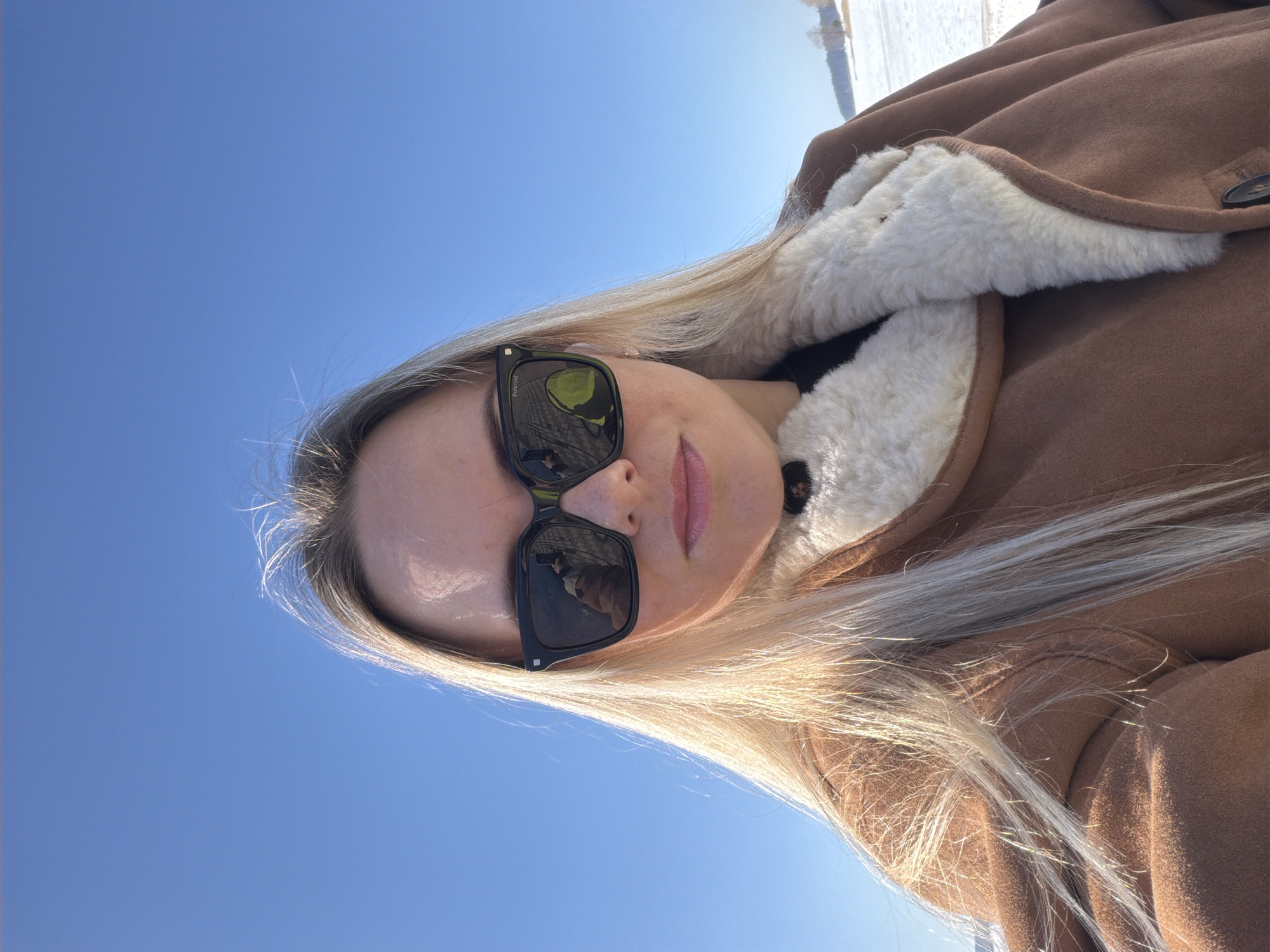 The image depicts a female individual taking a selfie outdoors in Votkinsk, Russia, under a clear, bright blue sky. The subject, positioned centrally, has long, straight blonde hair and wears black rectangular Polaroid sunglasses, which obscure her eyes. The reflections in the sunglasses show a person holding a camera (likely the subject herself) and another individual wearing a bright yellow-green coat. She is dressed in a brown or tan coat featuring a prominent cream-colored shearling or faux fur lining, with a dark garment visible underneath. Her lips are visible with a light pink tint. The background reveals an expansive, snow-covered landscape, consistent with a frozen body of water or field, extending to a distant treeline on a low hill. The scene is illuminated by strong, direct sunlight, indicative of a winter or early spring day.