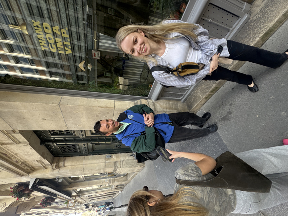 Three individuals are present on a narrow city sidewalk next to a stone-facade building on an unnamed street in an unknown city. On the left, a blonde female, seen from behind, wears a patterned t-shirt, grey sweatpants, and a dark brown suede tote bag. She holds a black smartphone horizontally. In the center, a male with short dark hair, wearing a blue and dark green jacket over a light green shirt and dark pants, is laughing with his hands clasped. To the right, a blonde female stands near a large storefront window, smiling. She wears a light-striped button-up shirt over a white tank top, black pants, and black tabi-style flat shoes, with a brown and black crossbody bag. The storefront window prominently displays yellow text reading "THANX GOD I'M A V.I.P." and reflects a modern glass building. An ornate, dark double door is situated between the male subject and the building's main facade, which also features window boxes containing red flowers. Other pedestrians are visible in the distance along the street.