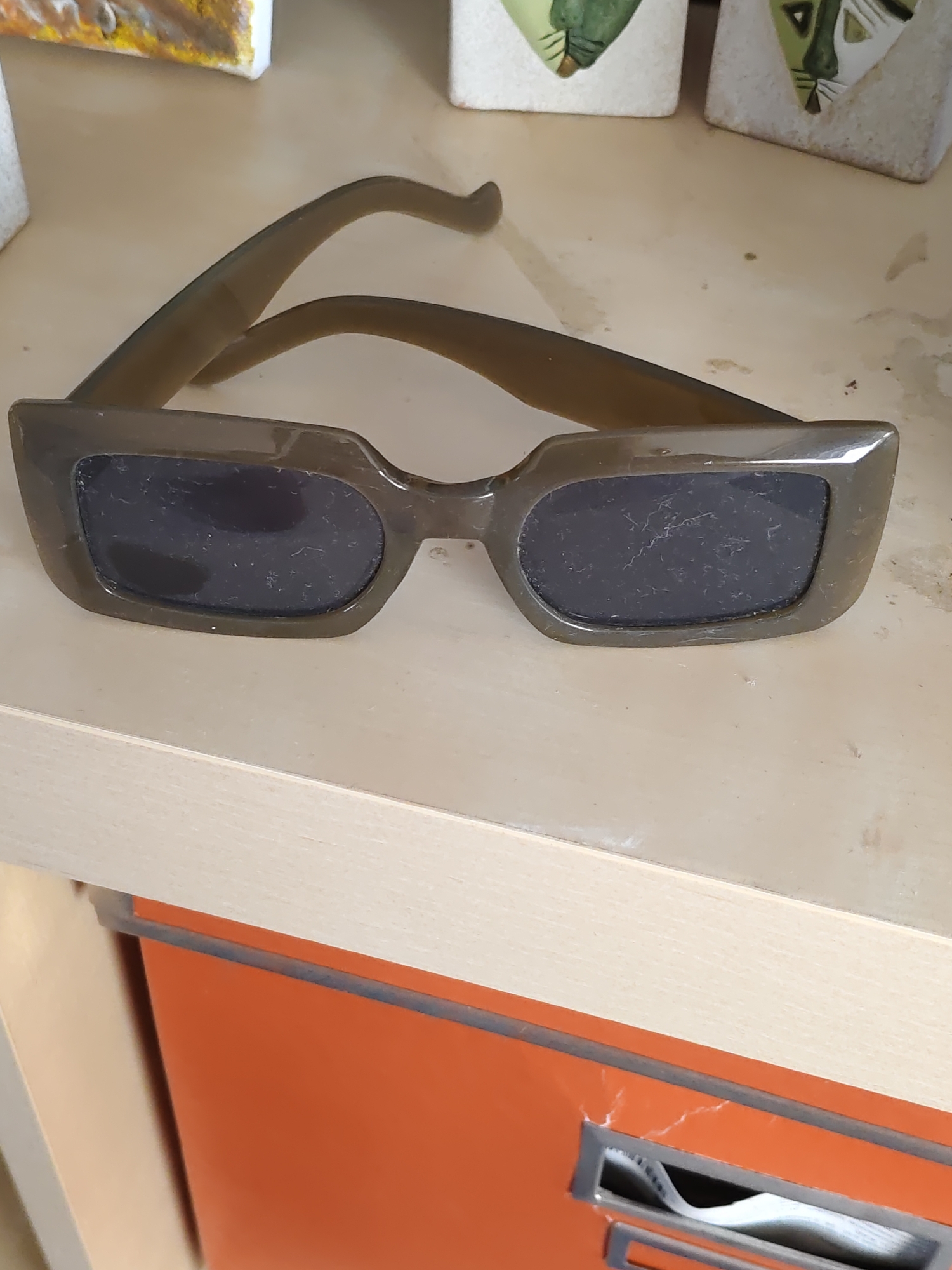 An overhead, close-up shot shows a pair of dark green, rectangular sunglasses with dark lenses resting on a light-colored, possibly wooden surface. The sunglasses are angled slightly, with the arms extending back and away from the viewer. In the background, out of focus, are three decorative objects, two of which appear to be ceramic blocks with stylized green and white animal faces. The surface the sunglasses are on is part of a shelf, with an orange drawer or cabinet visible below. The overall scene is indoors and appears to be a still life arrangement. The image location is specified as Rostov-on-Don, Russia.