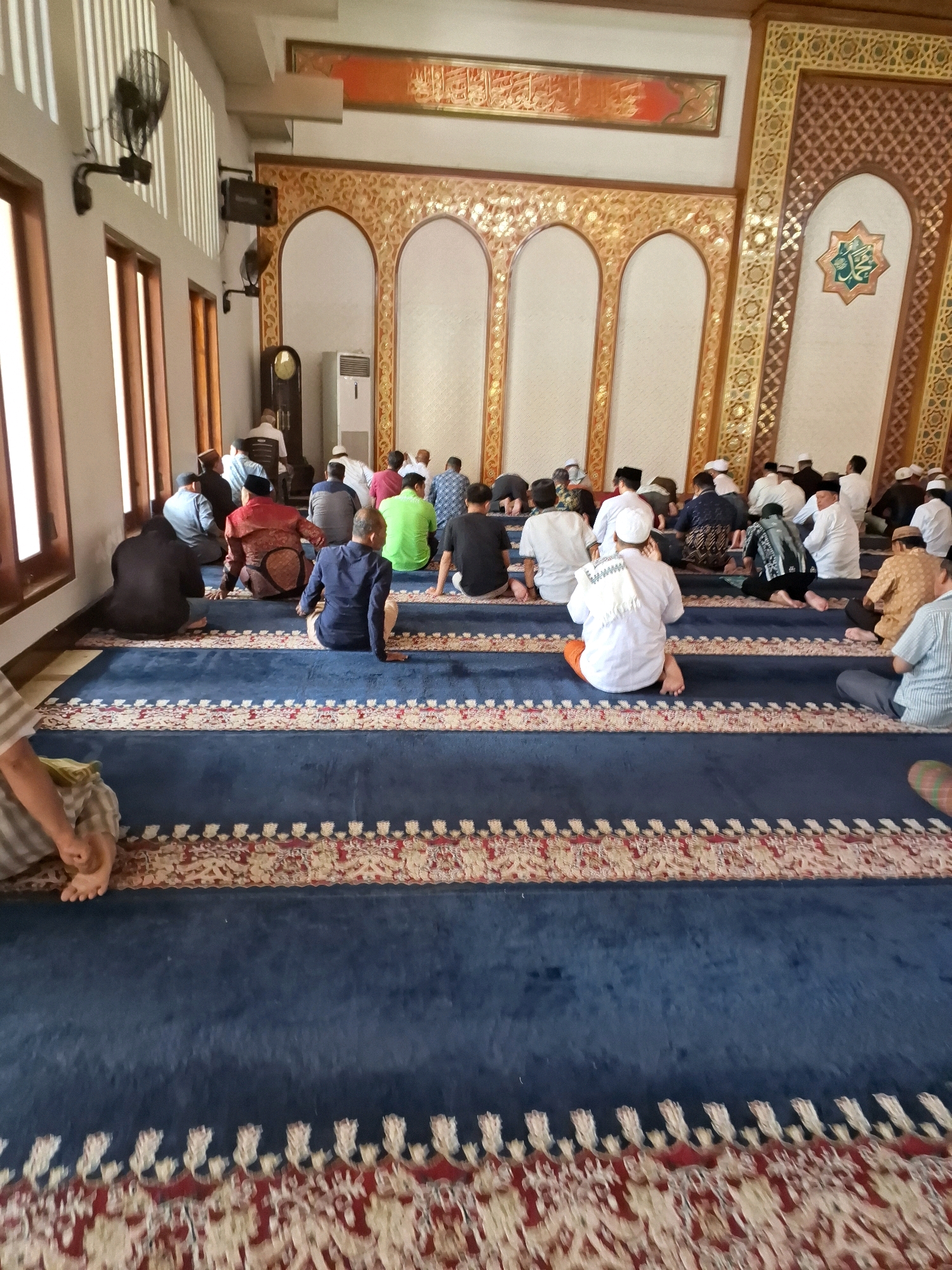 This image depicts a group of men engaged in prayer inside a mosque, likely in Gorontalo, Indonesia. The mosque features a high ceiling with intricate decorative elements, including arched doorways and ornate wall hangings. The men are seated on a large, blue carpet with red and white patterned borders, arranged in rows facing a decorated wall. Some men are wearing traditional Islamic attire, such as kufis and long robes, while others are in more casual clothing. The lighting appears to be natural, coming from windows on the left side of the image, and artificial lighting from fixtures on the ceiling. The overall atmosphere suggests a formal religious gathering.