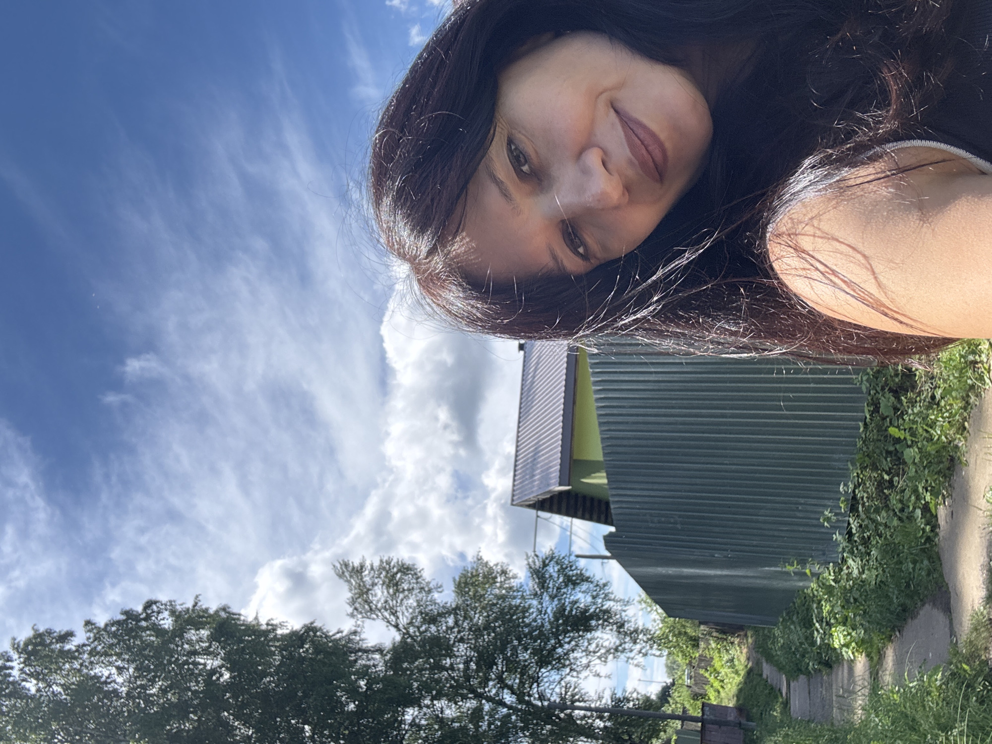 In an outdoor selfie taken in Chaikovsky, Russia, a woman with long, dark hair smiles gently at the camera. She is partially framed by her hair, with a portion of her bare shoulder visible. The background is a pastoral scene with a corrugated metal fence running horizontally across the middle ground. To the left of the fence, a green building with a dark metal roof is visible, nestled amongst trees. A narrow, unpaved pathway runs along the left side of the fence, bordered by overgrown green grass and foliage. The sky above is a bright blue, with scattered white clouds and wispy cirrus formations.