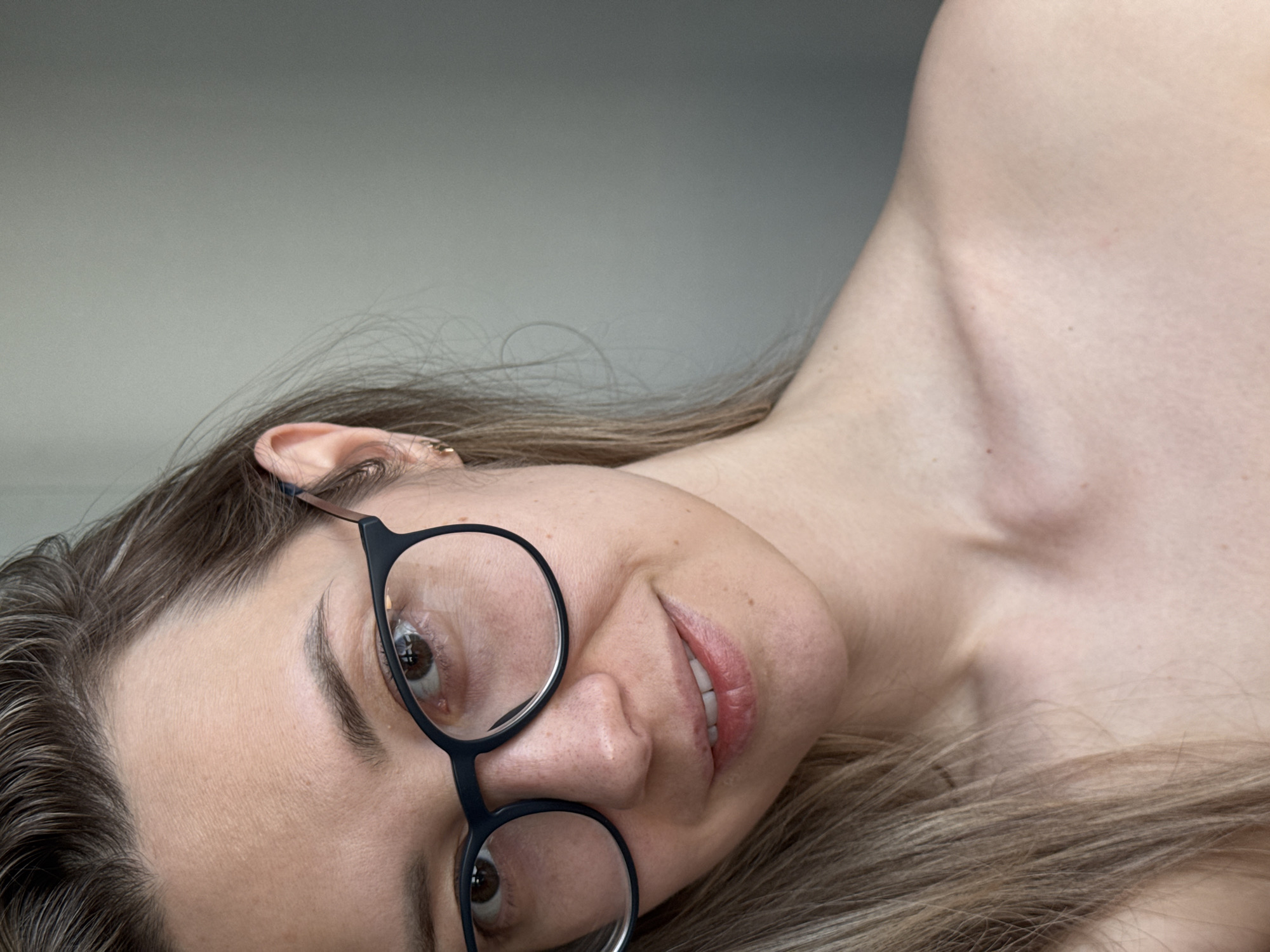 A fair-skinned woman with light brown hair is lying down, tilted on her left side. She is wearing black-rimmed glasses. Only a portion of her neck and shoulder are visible. There is a light gray background. The scene appears to be indoors. There is no clear indication that the image was taken in the city of Chaikovsky, Russia.