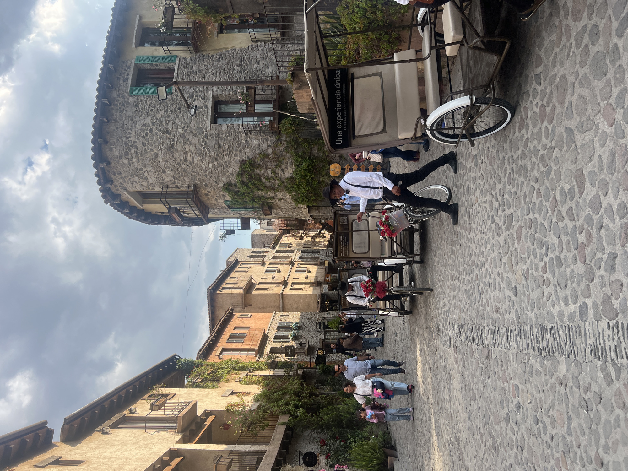 The image depicts a street in Domingo Arenas, Mexico, characterized by cobblestone paving and buildings constructed from stone and brick. Several individuals are present, including pedestrians and drivers of bicycle rickshaws. The rickshaws are adorned with flowers and advertisements, with drivers dressed in white shirts, black pants, and hats. The buildings exhibit varied architectural styles, with some featuring balconies and window boxes. The sky is overcast, suggesting cloudy weather.