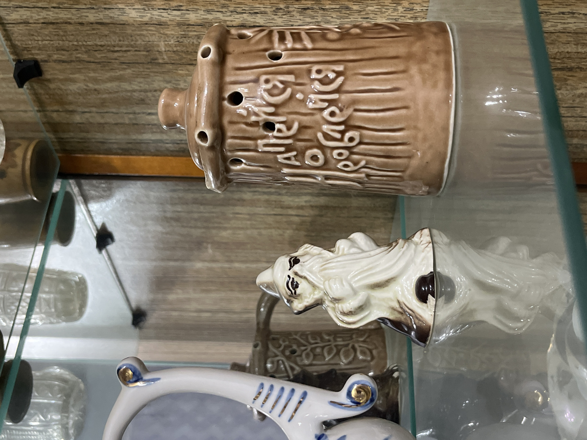 The image depicts a display case, possibly in a shop or private collection. Inside the case are several ceramic objects. A brown, cylindrical object with holes and stylized text is lying on its side. Below it is a white, decorative figurine that appears to be a mythical creature or deity, also lying on its side. In the foreground, a partially visible white and blue object, possibly a frame or ornate dish, adds to the collection of items within the case. The background is a light-colored wooden surface. It is unknown if this image was taken in the city of Chaikovsky, Russia.