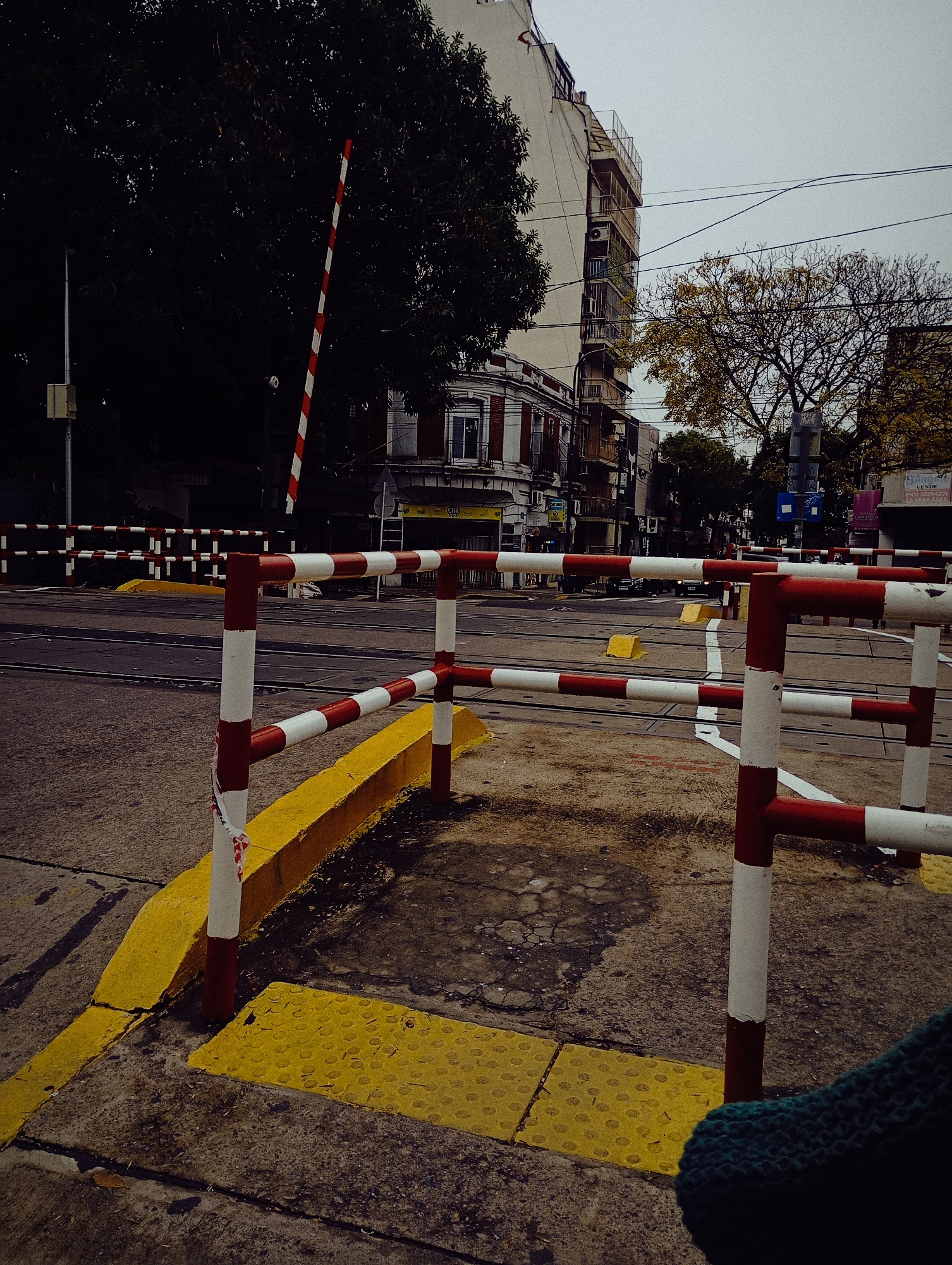 An eye-level shot depicts a railroad crossing in the city of Buenos Aires, Argentina. The foreground features a pedestrian barrier of red and white striped poles. The curb is painted bright yellow with a tactile paving surface, also yellow, designed for the visually impaired. The barrier runs parallel to the train tracks, which are visible in the middle ground. Across the tracks, buildings line the street. Trees with sparse foliage stand on either side of the street. A striped red and white crossing gate is raised on the left. The sky is overcast. A section of green fabric is partially visible in the lower right corner of the image.