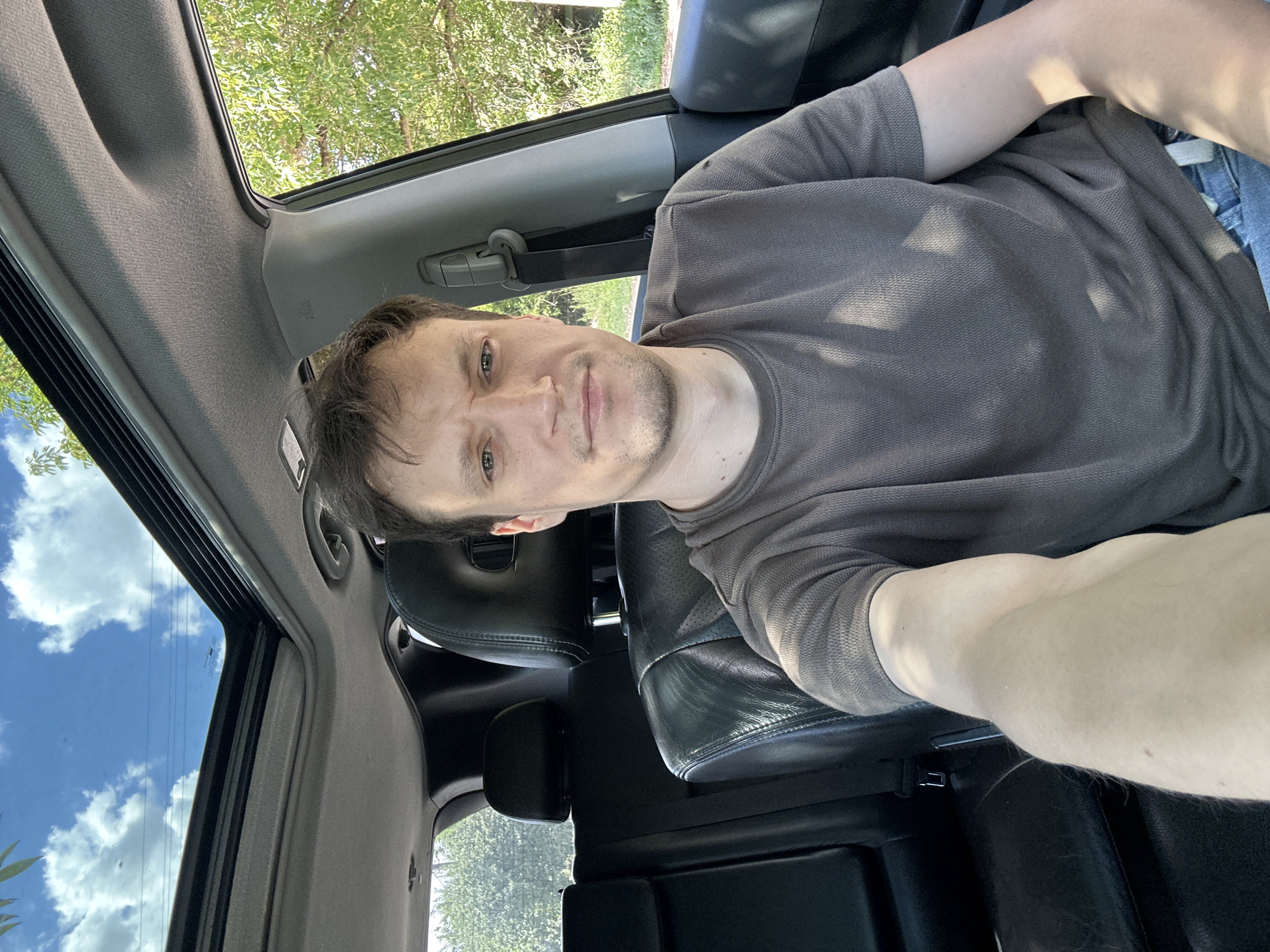 A man with short brown hair and a light beard sits in the driver's seat of a car, holding the camera to take a selfie. He is wearing a brown t-shirt and blue jeans. The car's sunroof is open, revealing a blue sky with white clouds. Trees and greenery are visible through the open sunroof and the car window. The interior of the car is dark, with black leather seats. The image appears to have been taken during the daytime, with natural lighting. The location is identified as Makiv, Ukraine.