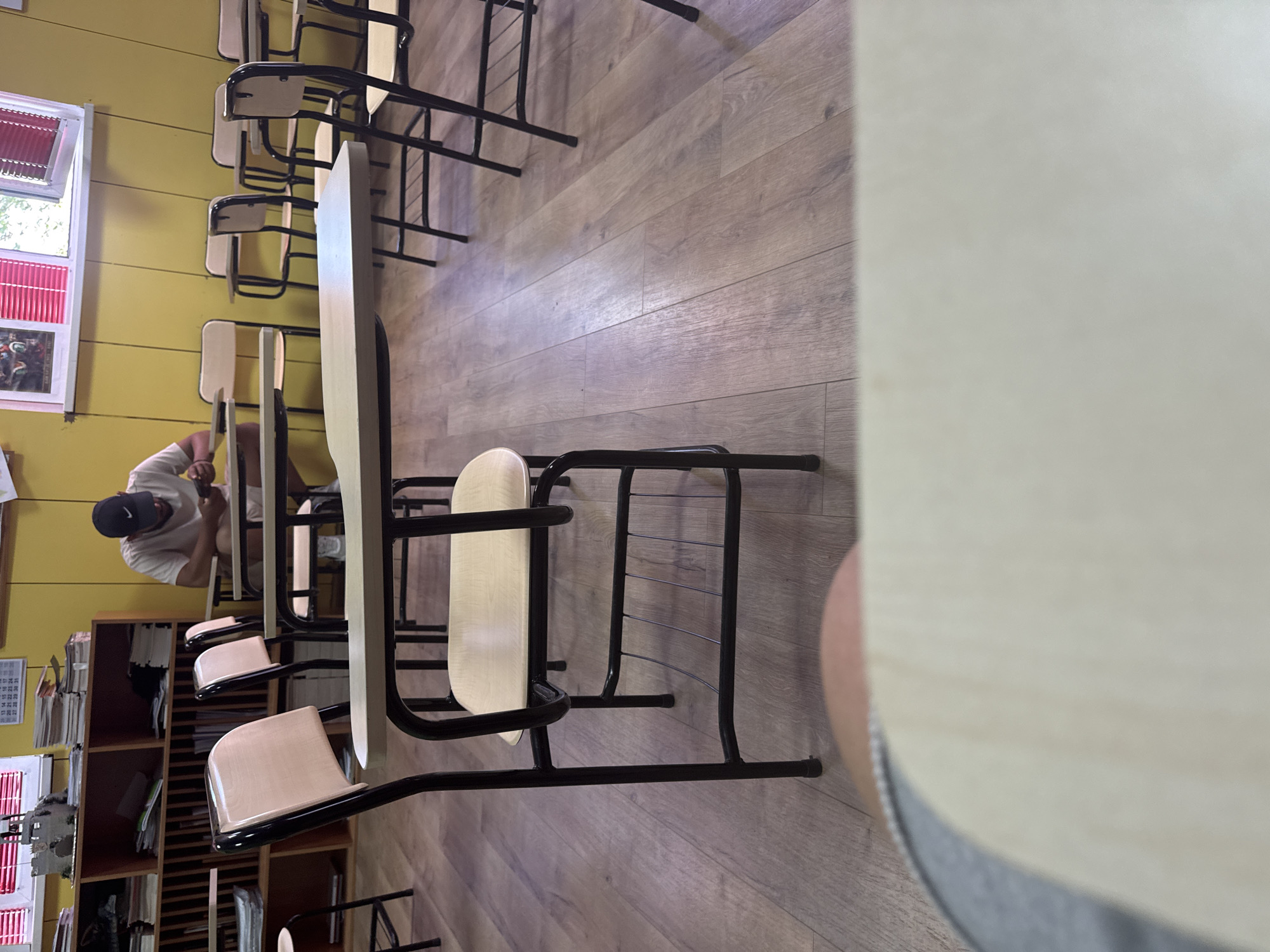 The image depicts a classroom with a wooden floor and yellow walls. In the background, a young man wearing a white t-shirt and a black baseball cap is seated at a desk, looking at his mobile phone. Several rows of desks with attached chairs are visible throughout the room. A shelf unit filled with books and papers is against the left wall. The image appears to be taken from the perspective of someone sitting at a desk in the foreground, with a portion of another desk and the observer's leg visible. The overall impression is that of an empty or sparsely populated classroom, with the focus on the student in the background and the arrangement of the furniture. There is no information within the image to suggest the location in Plovdiv, Bulgaria.