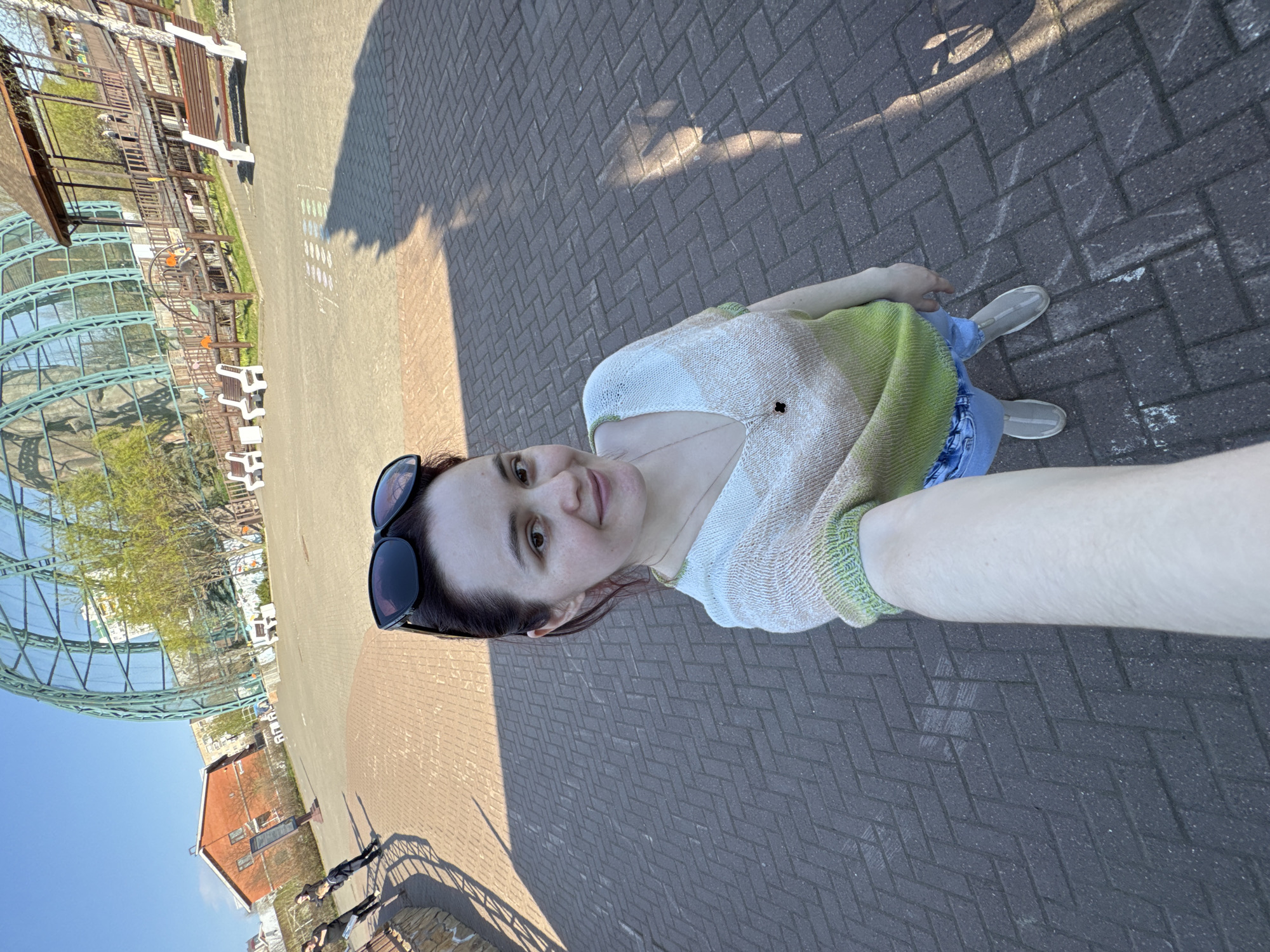 A woman is taking a selfie on a paved walkway in Izhevsk, Russia. She has dark hair and is wearing sunglasses, a light-colored top, and jeans. Behind her is a large greenhouse-like structure, benches, and a red brick building. The walkway is made of dark-colored bricks arranged in a herringbone pattern. Shadows are visible on the walkway. The weather appears sunny and bright.