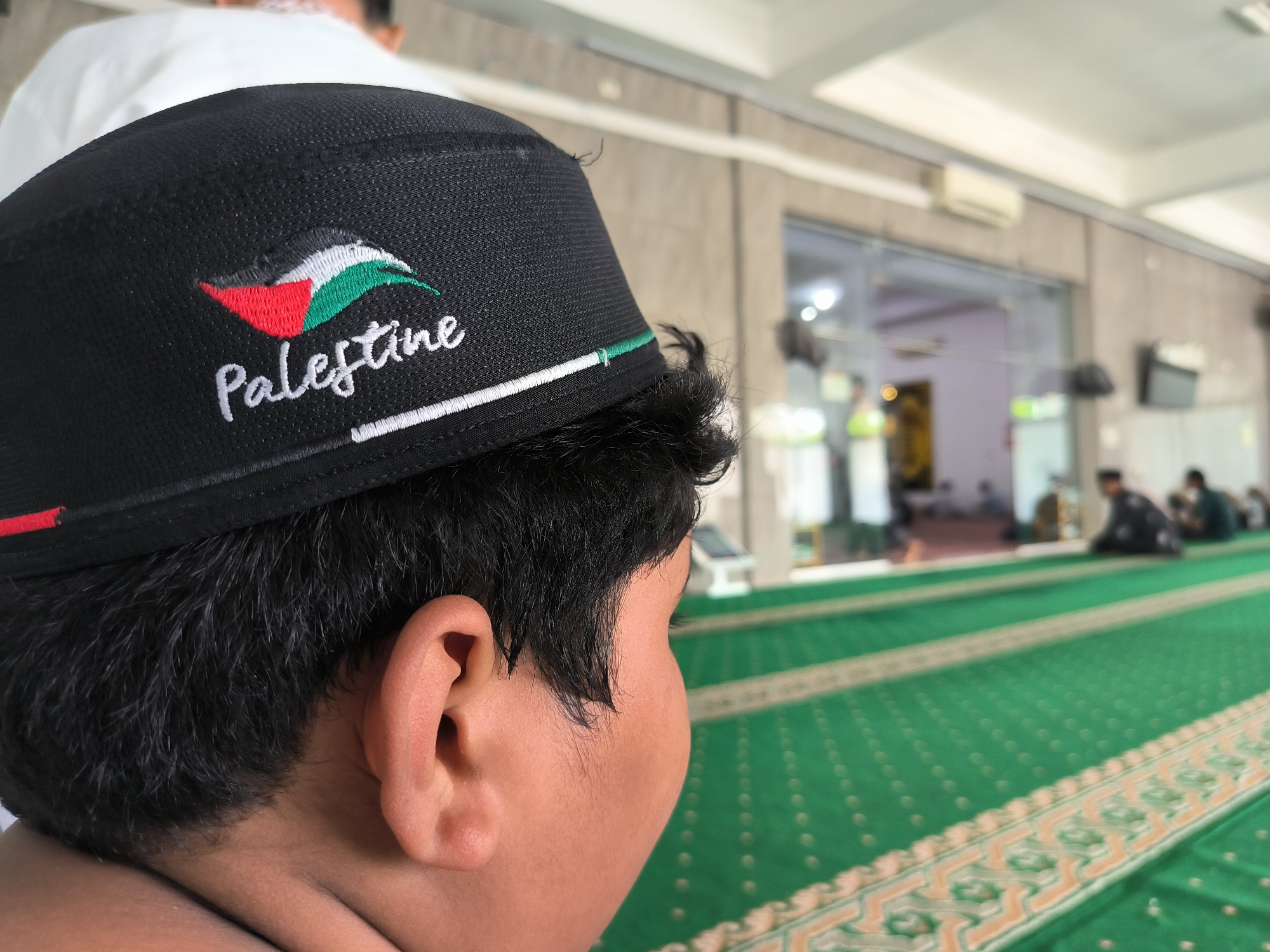 A child wearing a black cap adorned with the Palestinian flag and the word "Palestine" is pictured inside a mosque in Lhokseumawe, Indonesia. The mosque's green carpet is visible, and other individuals can be seen in the background. The child is facing away from the camera, showing their ear and the back of their head.