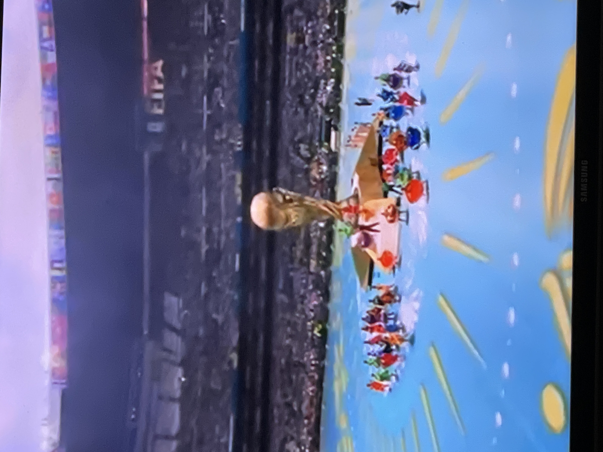 The image captures a stadium scene, likely during a sporting event. A prominent golden trophy, resembling the FIFA World Cup, stands on a raised platform in the center. Surrounding the trophy are numerous small figures, dressed in colorful attire, arranged in groups. The ground is a bright blue, adorned with yellow, elongated oval shapes. In the background, a large crowd fills the stadium seating, and the word "FIFA" is visible on a large screen. The lighting suggests an indoor or evening event. The stadium appears to be located in Moscow, Russia.