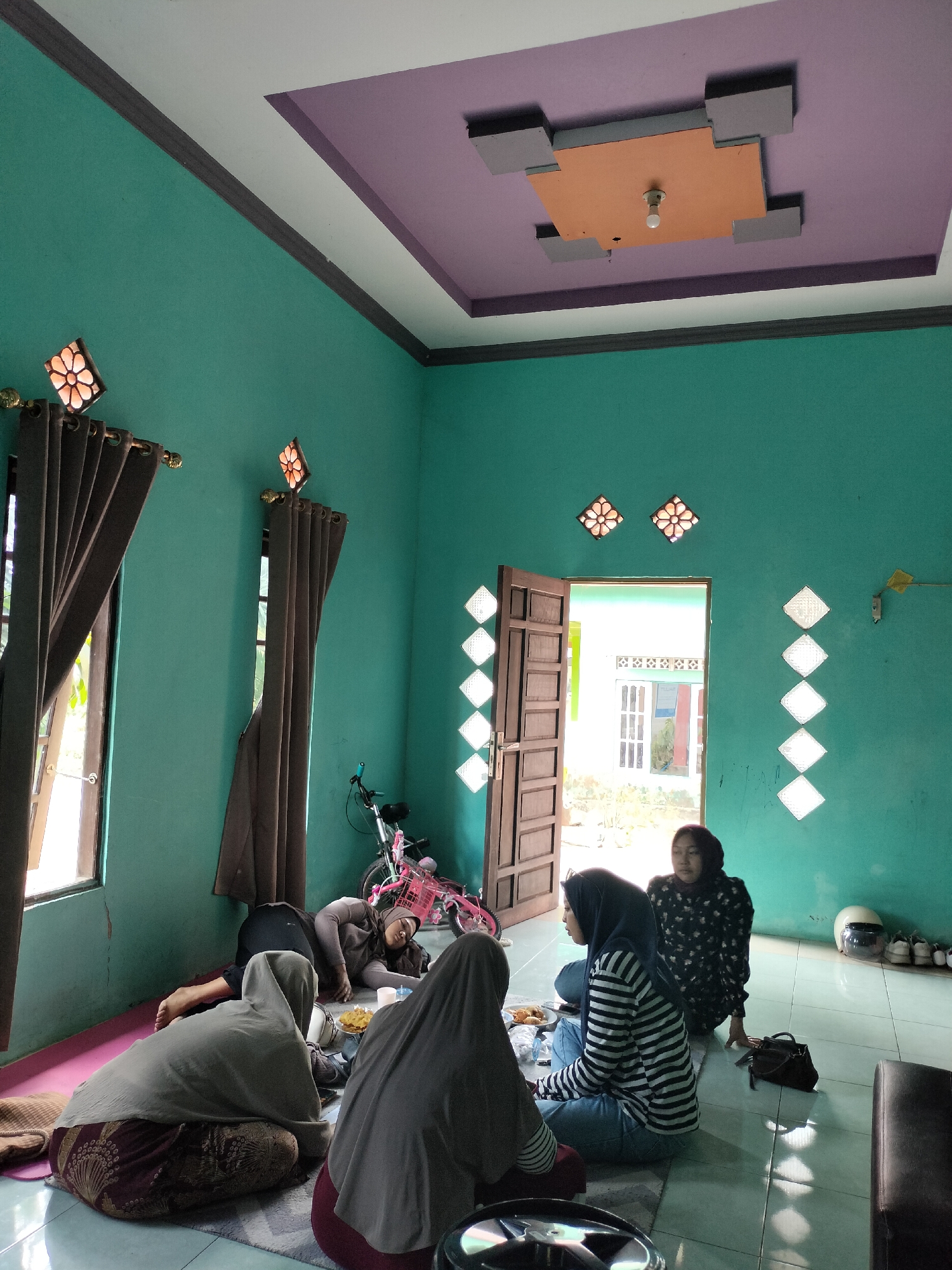 The image depicts an indoor scene with four individuals seated and lying on the floor, possibly in a living room or communal space. The room has teal-colored walls and a distinctive ceiling with purple and tan accents. The floor is tiled.
In the foreground, three individuals are seen from behind or the side. One person wears a grey hijab and a patterned skirt, seated cross-legged. Next to them, another person in a blue and white striped shirt and blue jeans is seated, wearing a dark blue hijab. A third person, to the far right and also wearing a grey hijab, is partially visible.
In the middle ground, a person is lying down, possibly asleep, on their side with their head resting on their arm. They are wearing a grey long-sleeved top and dark pants.
The environment includes several objects. A bicycle with a pink basket is positioned in the background near a doorway. Two windows with dark curtains are visible on the left side of the room. The doorway in the center opens to an outdoor area, revealing a building with windows. Decorative diamond-shaped vents are present on the walls. On a shelf in the background, a white helmet and several pairs of shoes are visible. A dark handbag is on the floor near one of the seated individuals.
The lighting suggests it is daytime, with sunlight streaming through the windows. The overall atmosphere appears relaxed, with individuals in comfortable positions. The image was captured in Angsana, Indonesia.