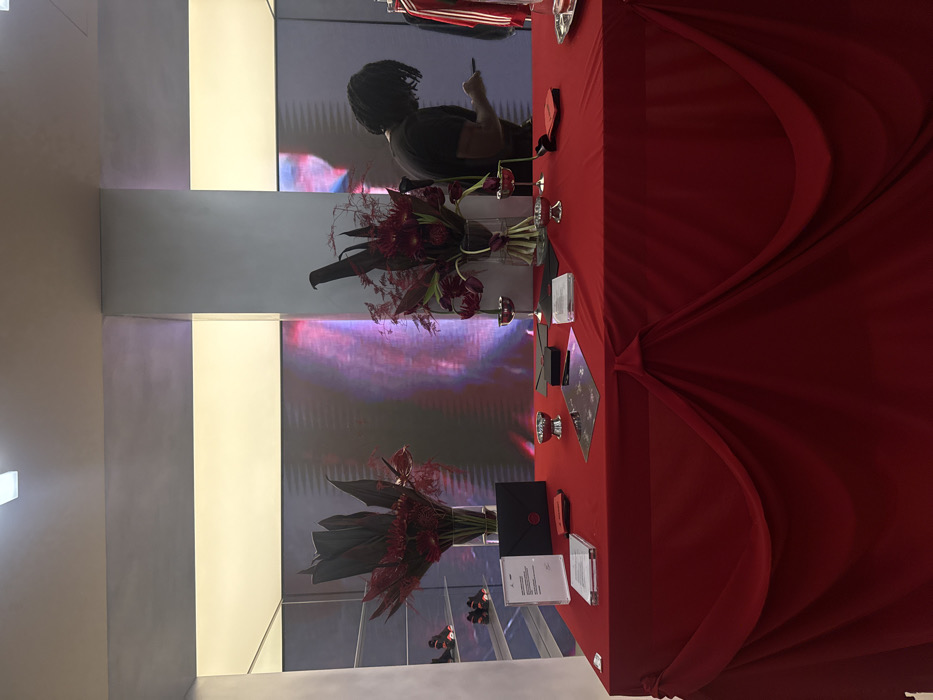 A red tablecloth draped over a table displays floral arrangements, envelopes, and mirrored coasters. A person stands near a video screen in the background. The scene features a modern architectural setting, possibly a store or event space, with a stark contrast between the clean lines of the architecture and the lush floral arrangements. The lighting highlights the table, creating a focal point in the composition.