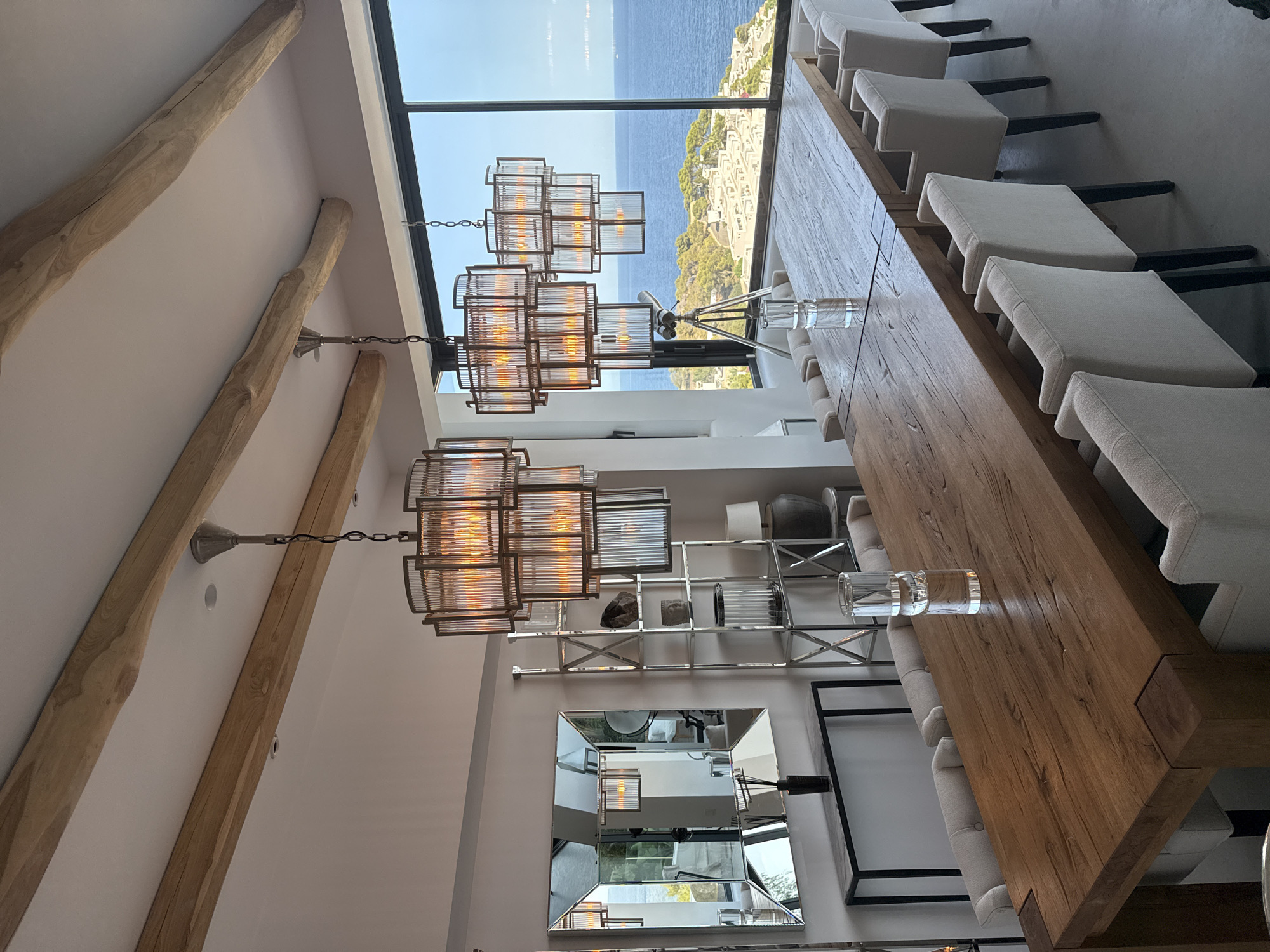 This is a photograph of a dining room with a large wooden table and chairs, set against a backdrop of a large window overlooking the sea. The room features exposed wooden beams on the ceiling and several modern glass and metal chandeliers. A large mirrored wall art is positioned on the left side of the room, reflecting the interior and the view. A chrome shelving unit with decorative items stands next to the mirror. The dining table is set with clear glasses. Outside the window, a view of the Mediterranean Sea and a coastal town can be seen. The scene suggests a contemporary, upscale dining area in a coastal property, possibly in Santa Eulària des Riu, Spain, given the architectural style and the prominent sea view. There are no people visible in the image.
