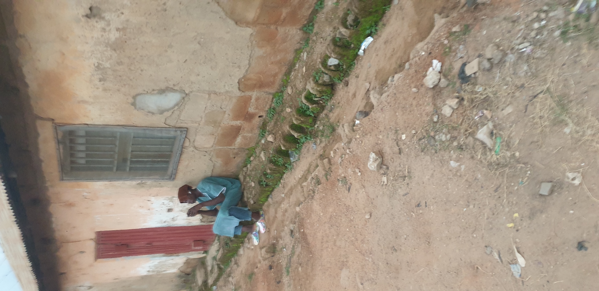 In Sabon Tasha, Nigeria, a lone individual, appearing to be an adult male, is seated on a raised concrete curb next to a weathered building. The man is wearing a turquoise colored tunic and dark trousers, with a brown head covering. He is holding a dark object, possibly a mobile phone, in his hands and appears to be looking down at it. The building behind him has a red corrugated metal door and a barred window. The ground in front of the building is a dirt path with scattered rocks and debris. A shallow, overgrown ditch runs along the curb where the man is seated, with moss-covered stones lining its edge. The overall scene depicts a moment of quiet repose in a somewhat disheveled urban environment.