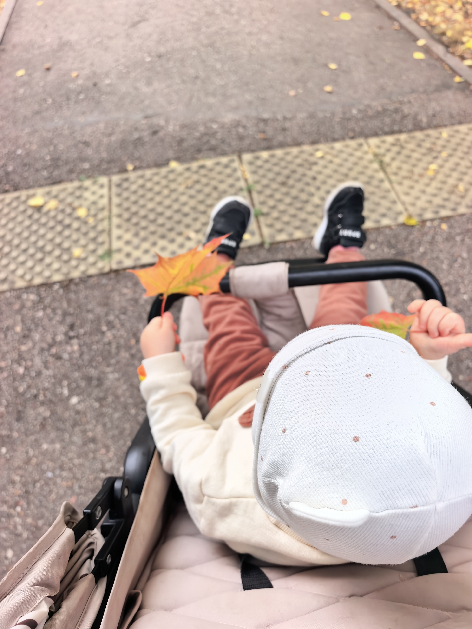 The image shows a close-up, overhead view of a young child sitting in a stroller. The child is wearing a white hooded jacket and rust-colored pants. A white hat with small pink dots covers the child's head. The child's left hand is visible, holding an orange and yellow maple leaf. The stroller is a light beige color with black metal accents. In the background, the ground appears to be a paved walkway with some fallen leaves. The context provided states the image is from Sterlitamak, Russia.