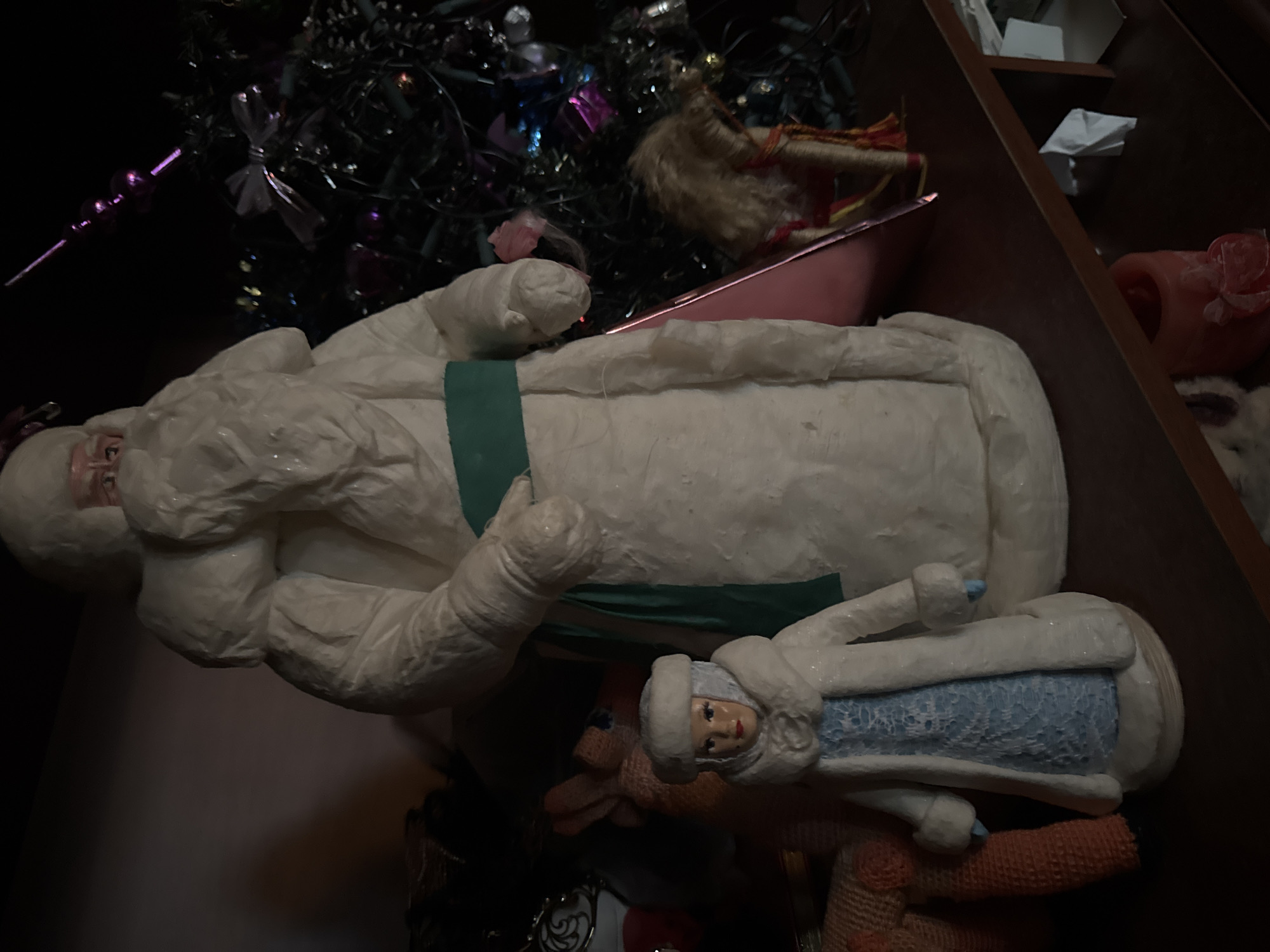 The image shows a collection of vintage New Year's figurines, likely from the Soviet era, displayed on a shelf. A large, white figure of Ded Moroz (Grandfather Frost) stands prominently in the center, wearing a traditional fur-lined coat and a green sash. To his left, a smaller figure of Snegurochka (the Snow Maiden) is visible, dressed in a blue and white outfit. Behind these figures, partially obscured by shadows, is a decorated Christmas tree with tinsel and colorful ornaments. Other objects on the shelf, such as stuffed animals and what appear to be additional figurines or decorative items, are also present. The lighting is dim, casting shadows and highlighting the textured surfaces of the figurines. The shelf itself appears to be made of dark wood. The overall impression is one of a nostalgic holiday display. The image was taken indoors, in a residential setting, possibly in Sertolovo, Russia, given the cultural context of the figurines.
