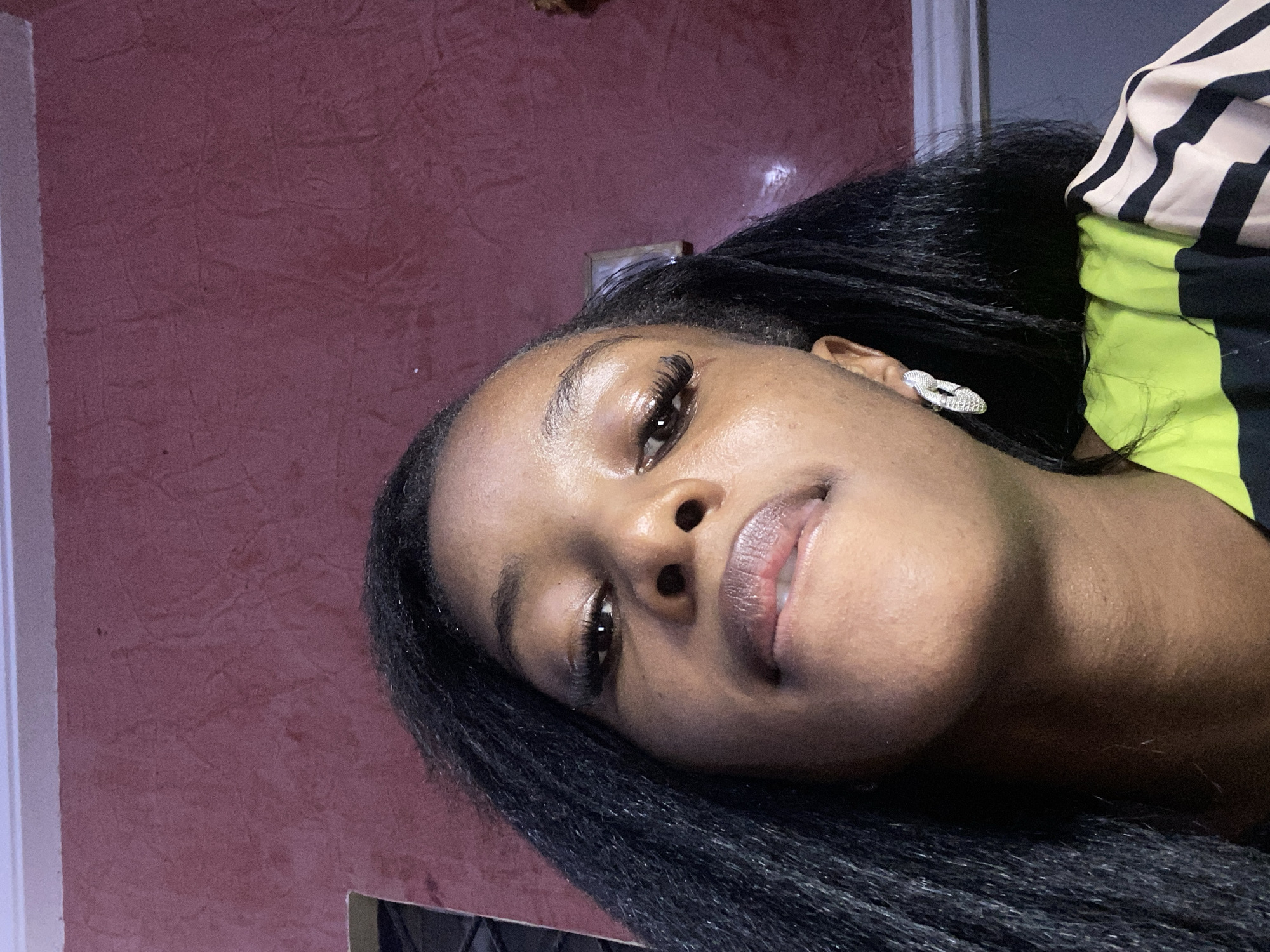 A young Black woman is captured in a low-angle, close-up selfie, her face tilted upwards towards the camera. She has long, dark hair that frames her face, and she is wearing a silver, intricately designed earring in her left ear. Her lips are slightly parted, with her tongue visible between them. Her makeup includes dramatic false eyelashes. She is wearing a shirt with a neon green and black striped pattern. The background is a textured, deep red wall, typical of many interior walls in homes and establishments in Jos, Nigeria. The lighting suggests an indoor setting.