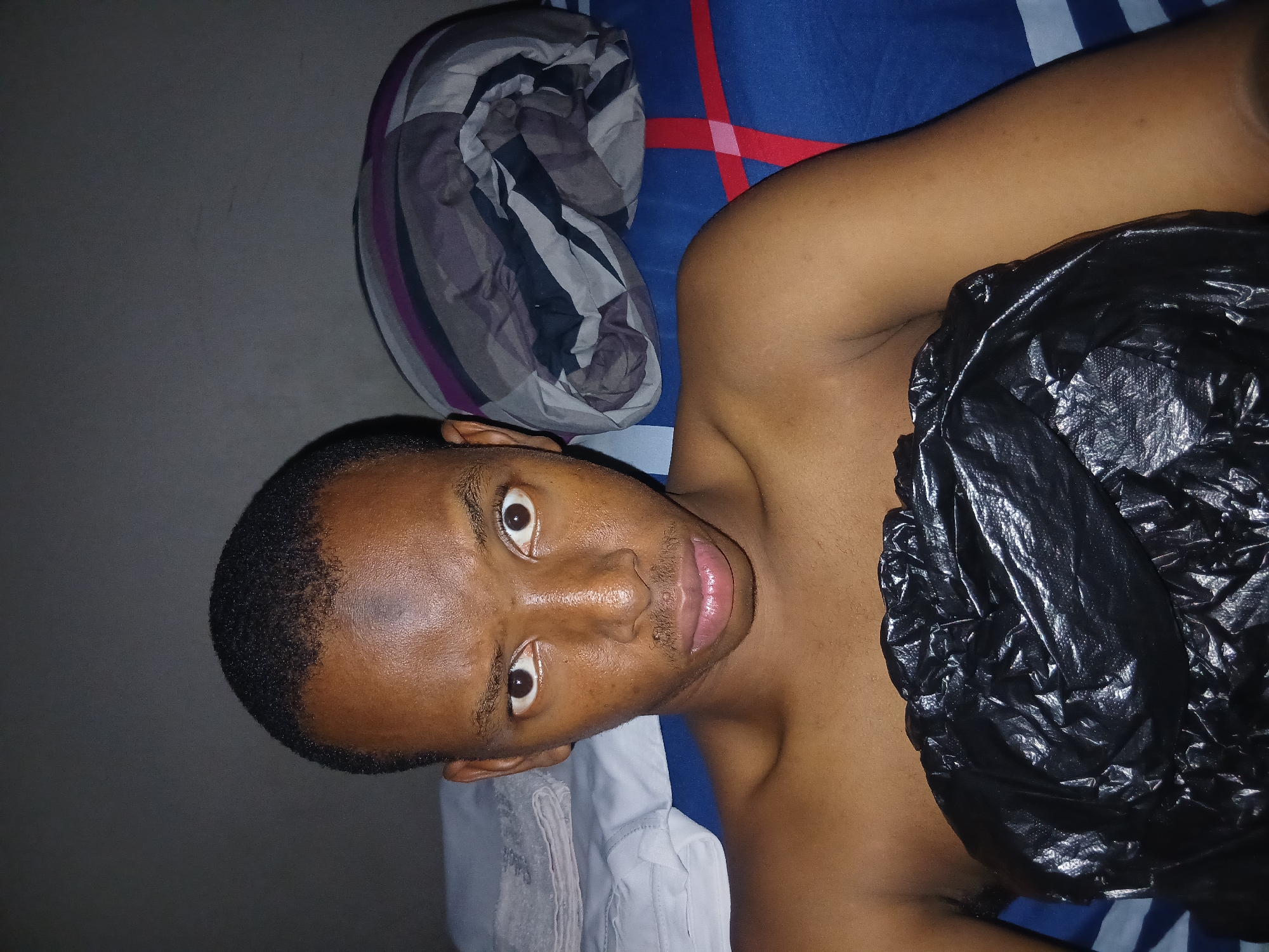 A young Black male is positioned centrally in the frame, looking directly at the camera with wide, curious eyes. He has a short, dark haircut and a faint mustache. His head is slightly tilted, and his expression is neutral. He is shirtless, with his upper body visible. He is wrapped in a large piece of crinkled black plastic, covering his chest and midsection.
Behind him, to the right, is a rolled-up blanket with a pattern of gray, purple, and black stripes. To the left of the frame, a folded white towel or sheet is visible, resting on a surface that appears to be a bed. The background is a dark, textured wall, suggesting an indoor setting. The bedding in the background has a blue base with red and white linear patterns. The overall lighting is dim, with the primary light source seemingly coming from above and in front of the subject, highlighting his face and the plastic wrapping. The environment suggests a domestic space, possibly a bedroom.