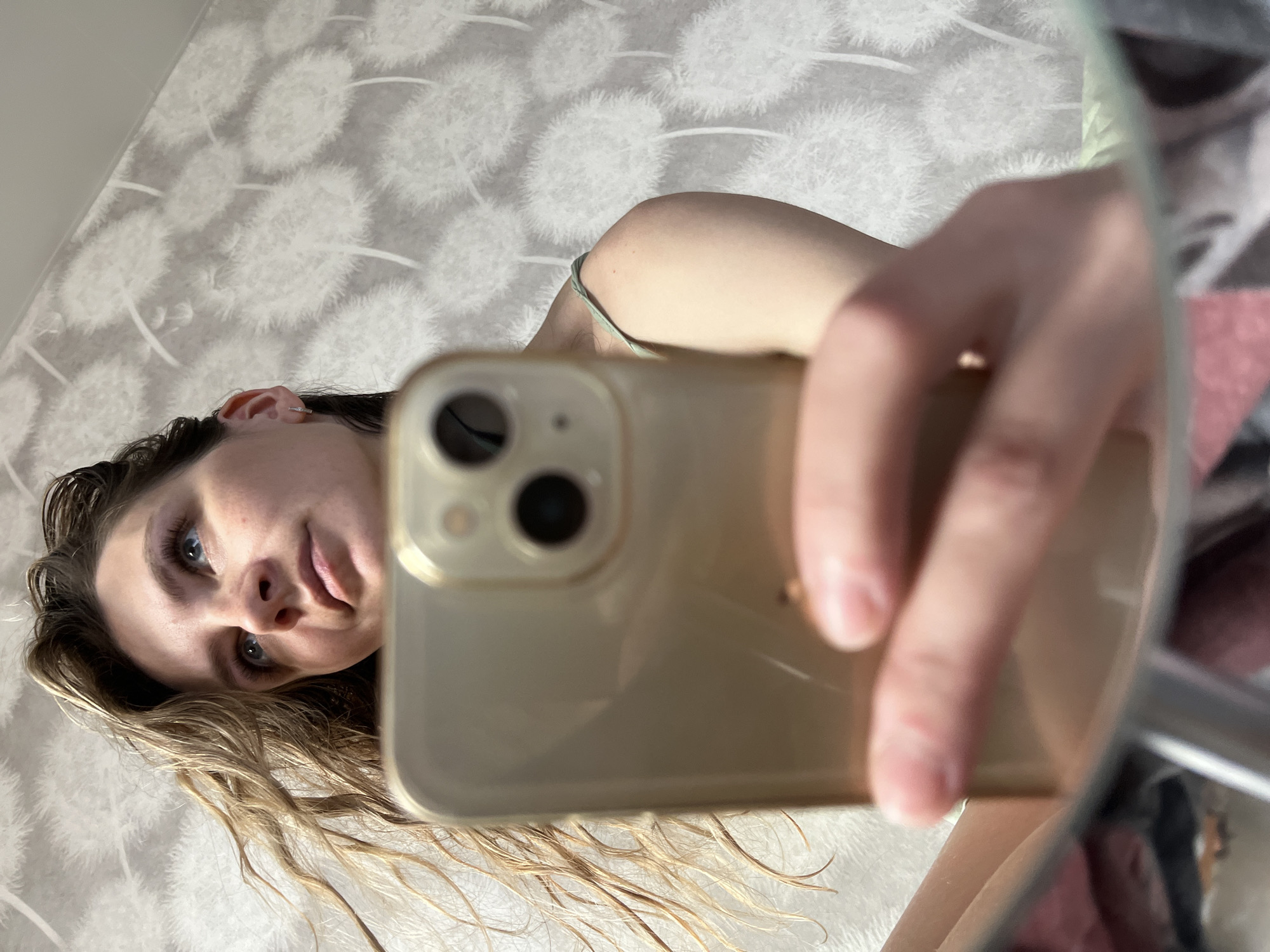 The image displays a mirror selfie of a young adult female in an indoor domestic setting. Her head and upper torso are visible, with her gaze directed upwards and to the viewer's right. She has light-colored, wavy hair, which appears damp, and blue eyes. Freckles are visible on her nose and cheeks. A small hoop earring is present in her left ear. She wears a light green or mint-colored spaghetti strap top. A gold-colored smartphone, identified by its visible dual-lens camera system, is held by her partially visible right hand, obscuring part of her lower face and left side. The background features a wall covered in light grey wallpaper patterned with numerous white dandelion seed heads. The bottom edge of the mirror and a blurred reflection of unseen domestic objects (possibly textiles in shades of pink, grey, and black) are visible at the very bottom. No specific location within the city of Chaikovsky, Russia, can be identified from the visual data provided.