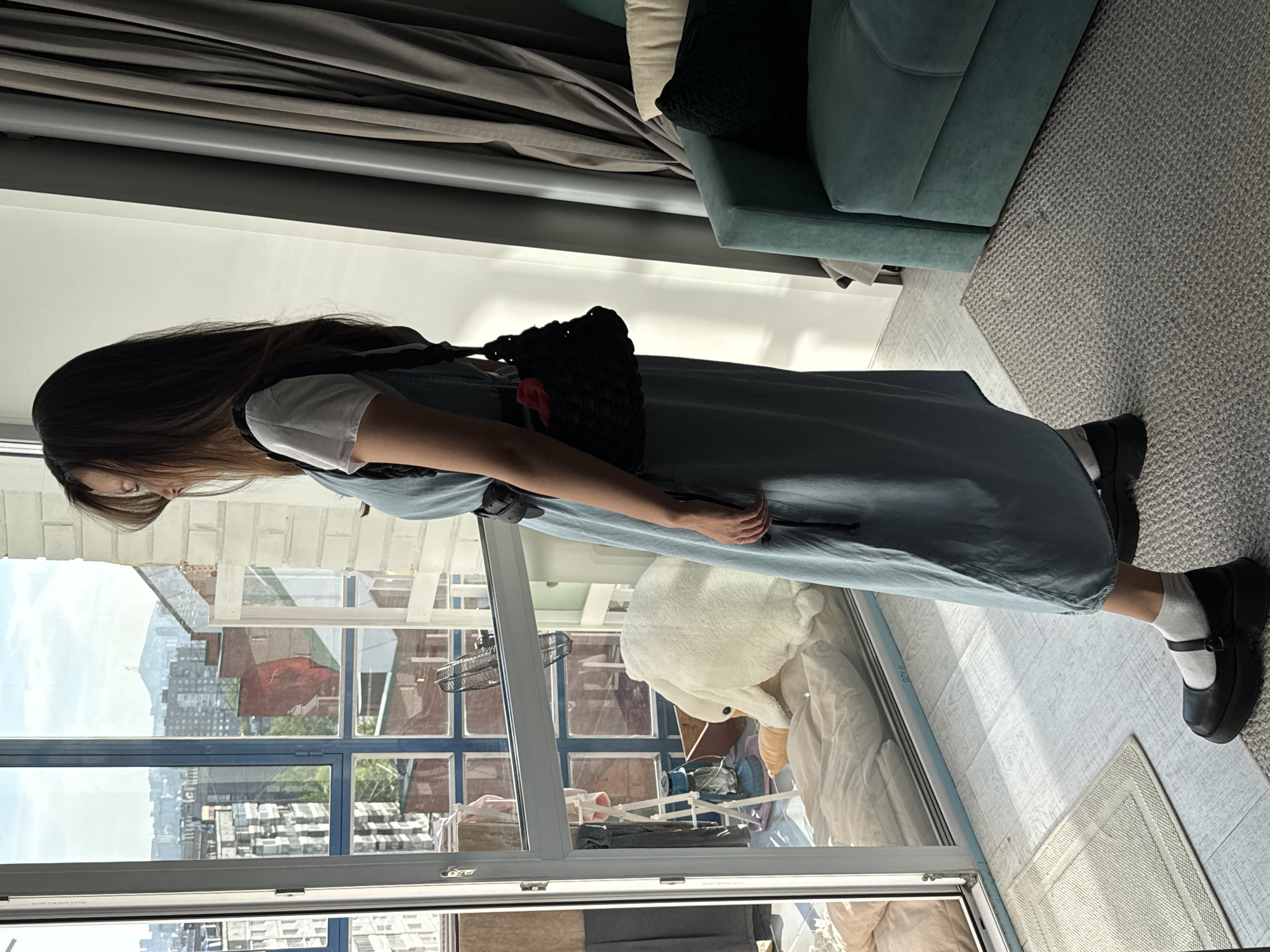 A young woman stands sideways, facing left, in an indoor setting with a large window. She has long, dark brown hair and is wearing a white short-sleeved t-shirt layered under a light blue, denim-like midi-length overall dress. A black woven shoulder bag with a subtle red accent hangs on her left shoulder. Her footwear consists of white socks and black platform Mary Jane-style shoes. She holds a dark, slim object, possibly a phone or remote, in her right hand.
The room features a teal-colored sofa with throw pillows (one dark green, one cream) and dark grey curtains. A light grey textured carpet covers part of the floor, transitioning to a light wood or laminate floor near the window.
Through the large window, a city skyline is visible under a bright sky, suggesting daytime. The view includes a variety of urban buildings, some red-bricked and others appearing as concrete apartment blocks. Distant hills or mountains can be seen on the horizon. On an exterior ledge or balcony just outside the window, a large white plush animal toy, resembling a swan or goose, is visible, along with a portable fan and a clothes drying rack with items on it. The bright natural light casts subtle shadows and reflections on the floor. The overall impression is a casual, relaxed moment in an urban apartment in Krasnoyarsk, Russia. No visible text is present.