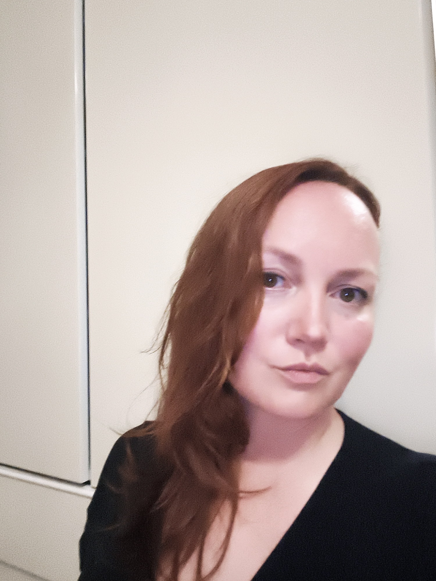 A woman with long, reddish-brown hair and fair skin is captured in a selfie. She is looking directly at the camera with a neutral expression. Her hair is parted to her left, with a portion falling over her left shoulder. She is wearing a dark-colored top that appears to be a V-neck. The background is a plain, light-colored wall, possibly the interior of a room, with a vertical line or edge on the left side, suggesting a corner or a doorframe. The lighting is soft and diffused, indicating it might be indoors during the day. There is no visible text in the image. The overall mood is calm and straightforward.