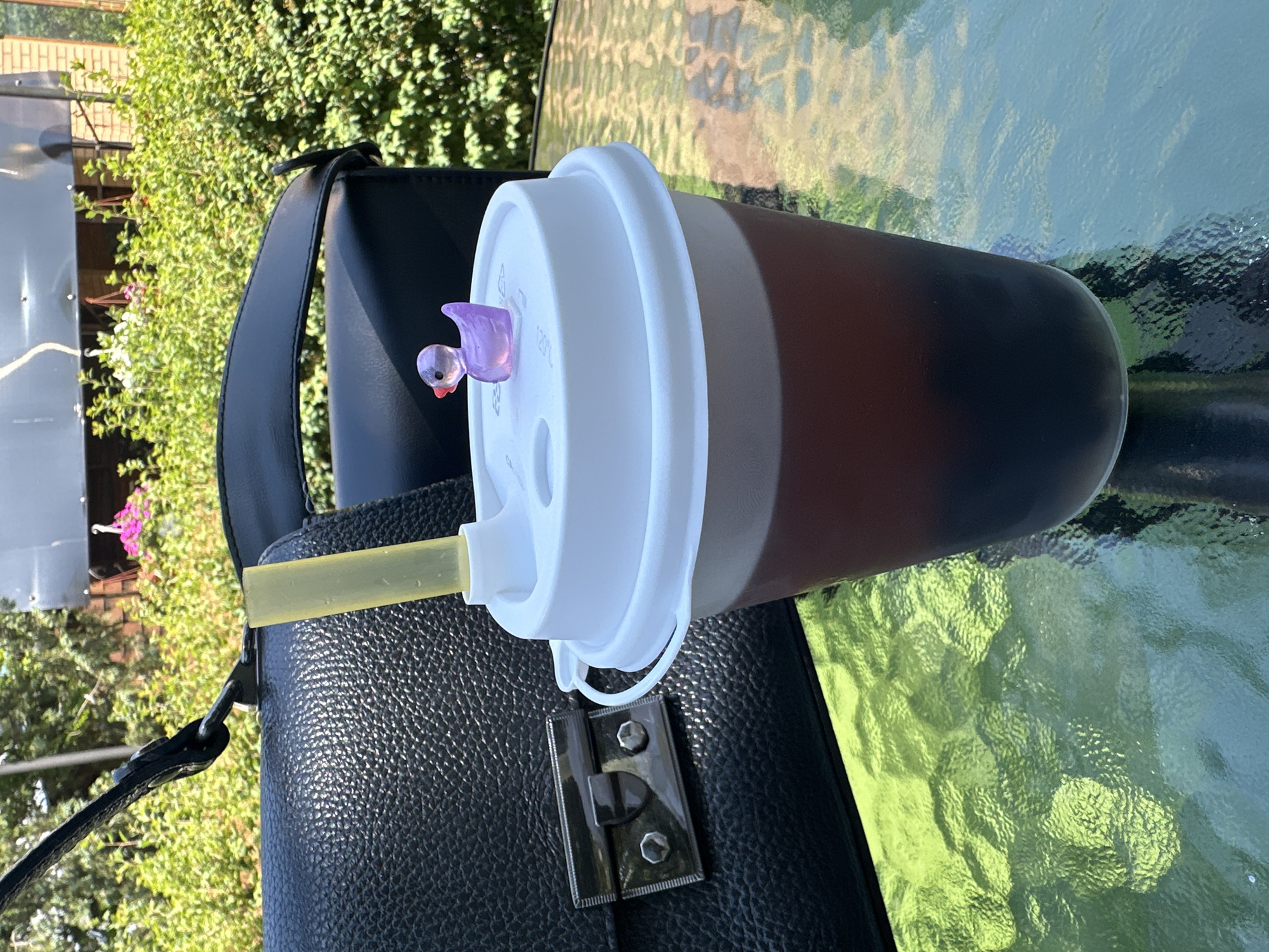 On a textured glass tabletop in Horishni Plavni, Ukraine, sits a disposable cup filled with a dark beverage, topped with a white lid and a yellow straw. A small, translucent purple duck charm rests on the lid. To the left of the cup, a black leather handbag with a silver clasp is partially visible. The background consists of lush green foliage and hints of other outdoor elements, suggesting an exterior setting. The lighting indicates it is daytime, likely a sunny day given the bright reflections on the table.