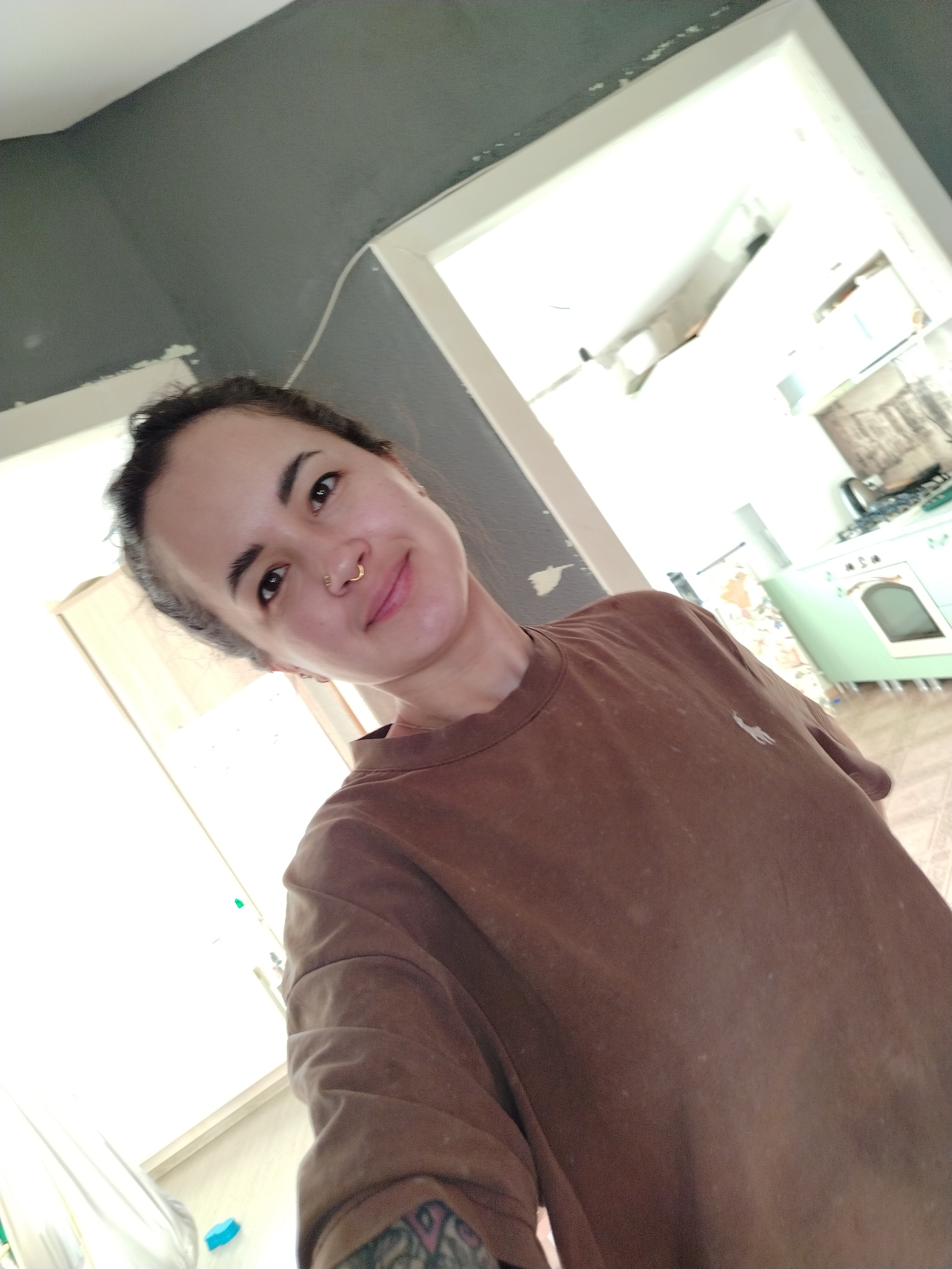 A selfie shows a woman with brown hair pulled back, a nose ring, and a brown t-shirt. She is standing indoors, with a doorway and a grey wall behind her. Through the doorway, a kitchen is visible with a light green oven and stove. The location is not discernible from this image.